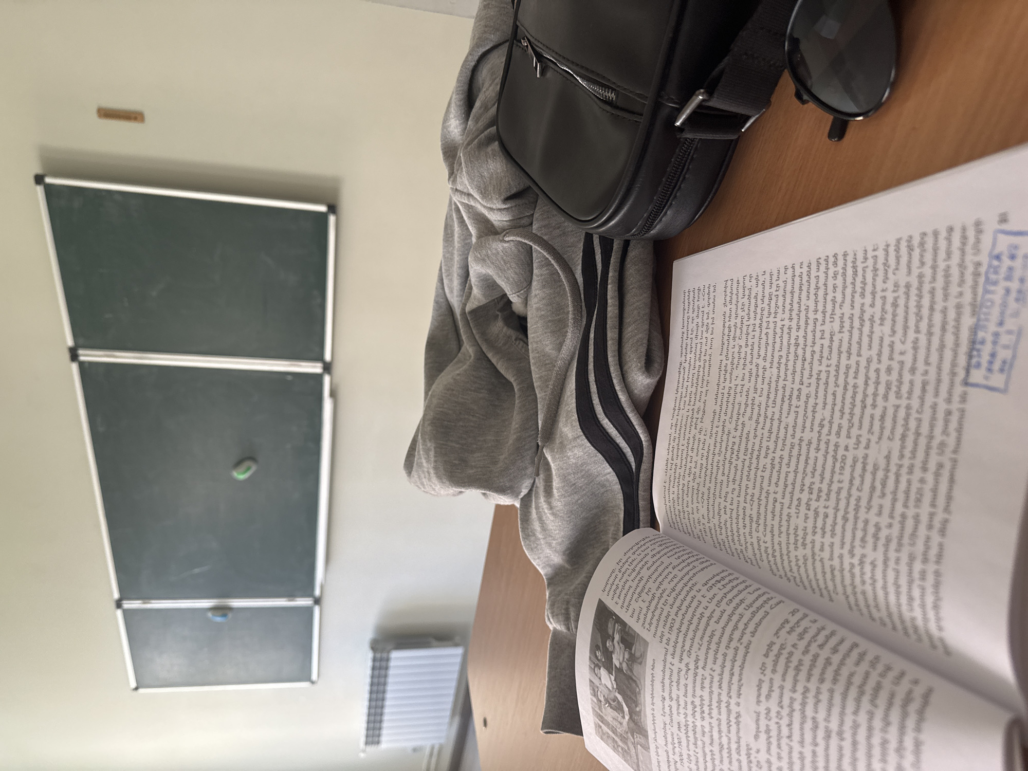 The image captures a scene indoors, seemingly on a desk or table. A book is open, displaying text primarily in what appears to be Armenian script. Adjacent to the book is a gray hoodie with dark stripes on the sleeves. A black shoulder bag rests next to the hoodie, with a pair of sunglasses on top of it. In the background, a chalkboard is visible on the wall, consisting of three sections stacked vertically. The presence of Armenian text suggests the location is likely Yerevan, Armenia. The scene depicts a typical study environment.