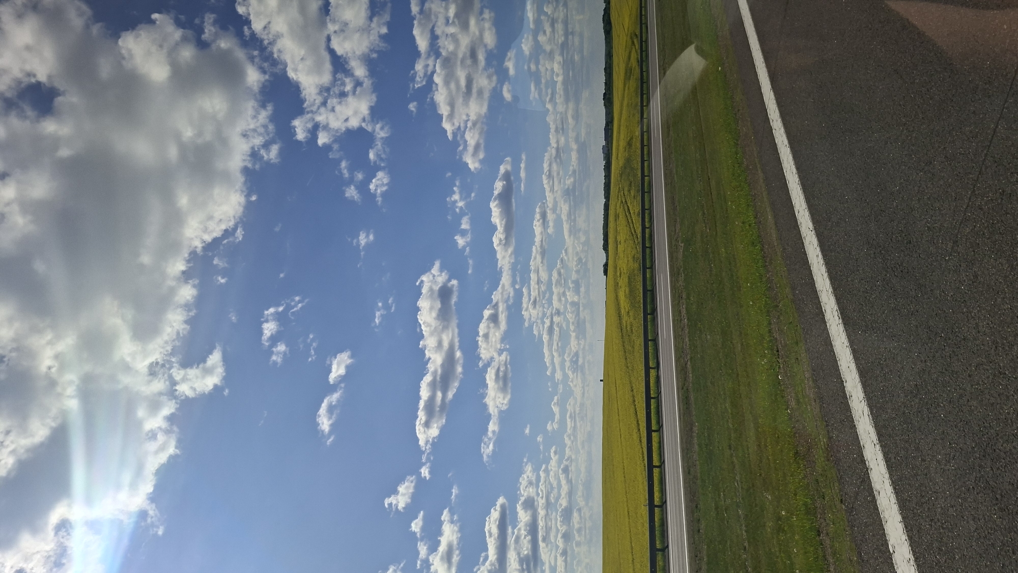 The image shows a view from a moving vehicle. The sky is blue with scattered white clouds, some of which have sunbeams shining through them. A road runs along the right side of the image, with a white line marking the edge. To the left of the road is a green field and a yellow field, separated by a guardrail. The location is Ozyaritska-Slabadski rural council, Belarus.