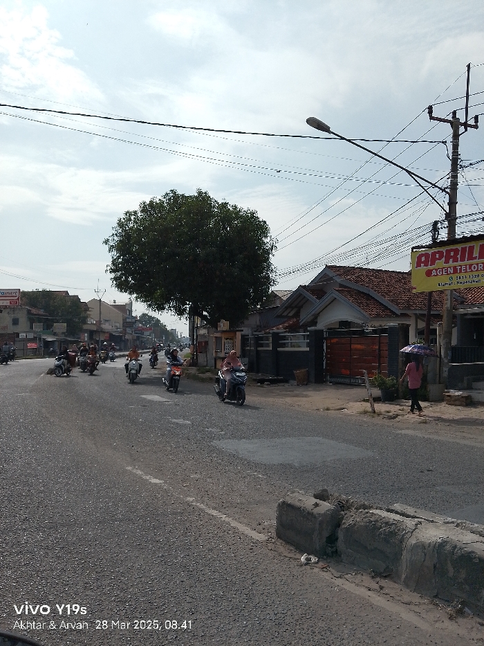 The image shows a street in Jatibarang, Indonesia, located at latitude -6.475, longitude 108.309. The scene is a street with a variety of buildings and businesses on either side. There are a few people riding motorcycles on the road. There is a person walking on the sidewalk and a sign for a business that says "APRIL" in large letters. There is also a tree in the middle of the road. The sky is clear and there are many power lines running overhead. The street is paved with asphalt and has some cracks and potholes. There are two concrete barriers on the right side of the road. A street sign is on the left side of the road. A person is holding a red umbrella and is walking in the sidewalk.