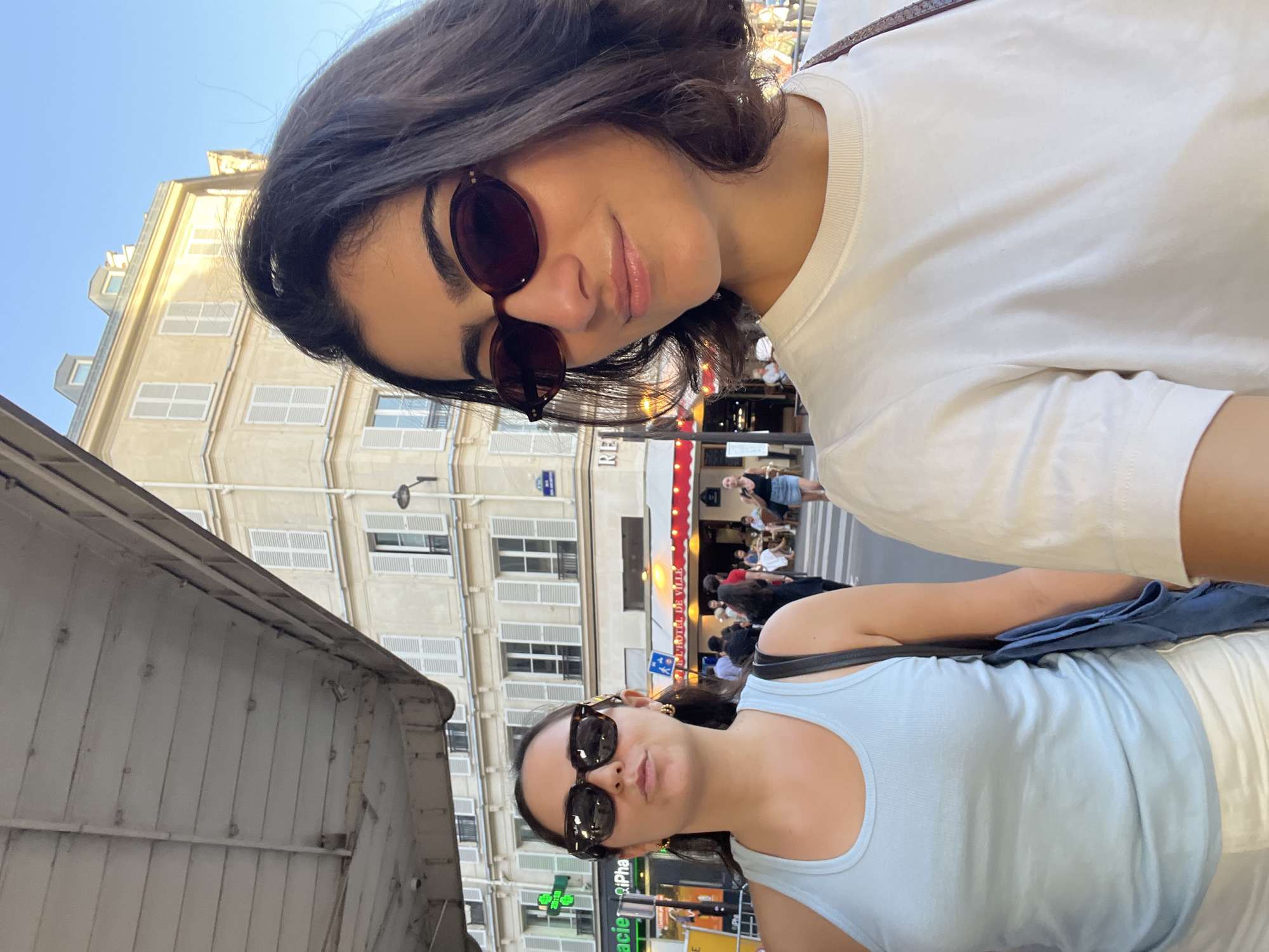 The image depicts two young women in an outdoor setting, likely a street in Paris, France. One woman wears a white t-shirt and sunglasses, while the other wears a light blue tank top and sunglasses. The background features Parisian architecture, including buildings with multiple windows and shutters. A street sign is partially visible, along with what appears to be a restaurant or café, with patrons seated outside under a red awning. The overall scene appears to be a sunny day in a bustling urban environment. There is a pharmacy as well in the picture.