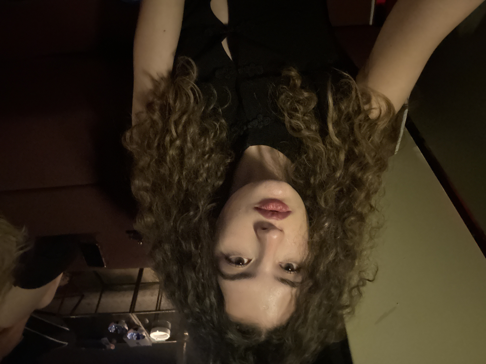 A young woman with curly brown hair and fair skin is looking directly at the camera. She is wearing a black top with ornate button closures and a V-shaped cutout. Her lips are painted with a deep red lipstick. She appears to be indoors, in a dimly lit environment. In the background, out of focus, a dark brown sofa is visible, along with a glass-topped table on which an ashtray and other objects are placed. The overall impression is of a casual indoor setting, possibly a lounge or bar. There is no indication of Milan, Italy, in the image.