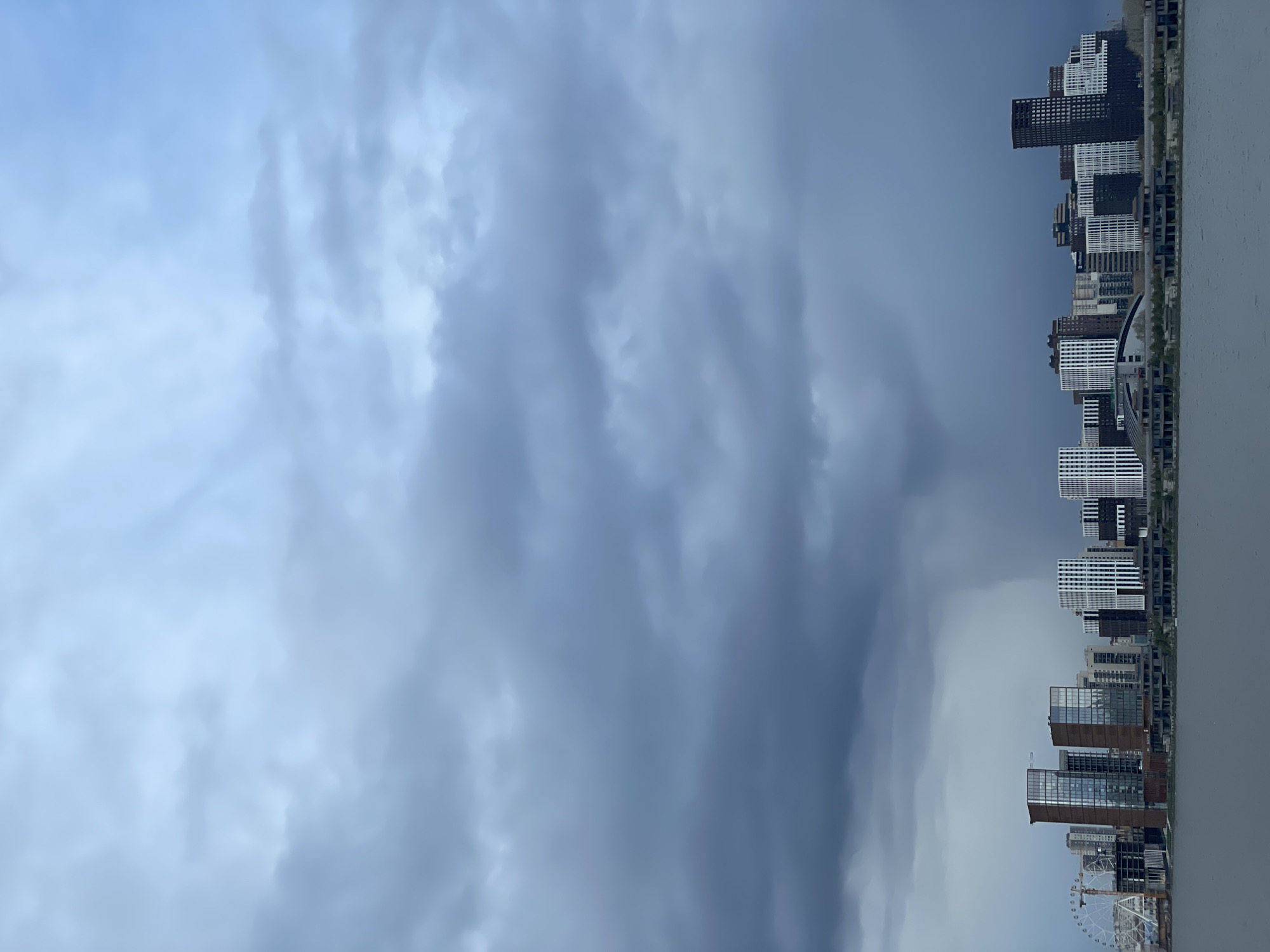 The image captures a panoramic view of a modern cityscape situated alongside a calm body of water, all set beneath a dramatic and heavily overcast sky. The scene is dominated by a vast expanse of clouds, occupying roughly two-thirds of the frame, with the city and water occupying the lower portion.
The sky is filled with dense, layered clouds exhibiting a range of grey tones, from lighter, diffused grey-blue on the left to darker, more turbulent and imposing grey masses towards the right. These heavy clouds suggest a heavily overcast day, potentially signaling an approaching storm or rainy weather, casting a subdued light over the entire landscape.
Below the cloud line, a dense array of contemporary buildings forms the city skyline. The structures are primarily high-rise, displaying diverse architectural styles with facades ranging from dark to stark white, and some featuring reflective glass. The buildings are tightly packed, indicating a bustling urban environment. On the far right side of the city's edge, a large Ferris wheel is visible, suggesting a waterfront entertainment or recreational area.
A serene, monochromatic body of water stretches across the foreground, reflecting the muted greys of the sky and visually separating the viewer from the distant city. No people or specific activities are discernible in the image. The time of day is daytime, but the thick cloud cover creates an atmospheric and somewhat dim lighting condition. No visible text or specific geographical identifiers are present, yet the architecture and scale suggest a major metropolitan area. The overall impression is one of quiet grandeur, marked by the powerful presence of the weather.