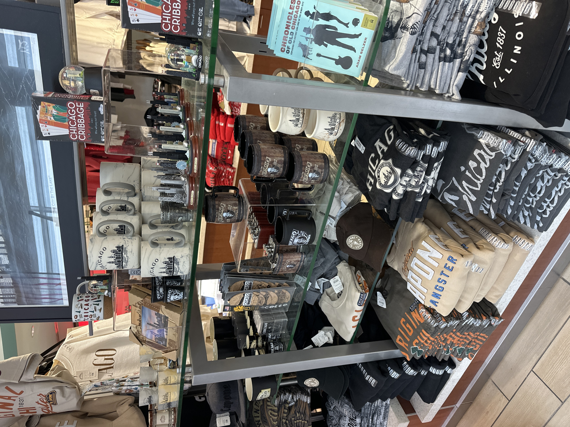 The image shows a retail display of Chicago-themed merchandise. The display is organized on multiple shelves, showcasing a variety of items such as mugs, shot glasses, snow globes, t-shirts, sweatshirts, and baseball caps. Many of the items feature iconic Chicago landmarks like the skyline, as well as references to the city's history and culture, such as "Windy City" and "Al Capone." There are also boxes of a game called "Chicago Cribbage." The merchandise is arranged in a neat and orderly fashion, typical of a souvenir shop or gift store. The background suggests an indoor setting, possibly a store or museum gift shop. The lighting is bright and even, illuminating the products clearly. There are no people visible in the image. The overall impression is one of a well-stocked retail environment selling souvenirs related to Chicago.
