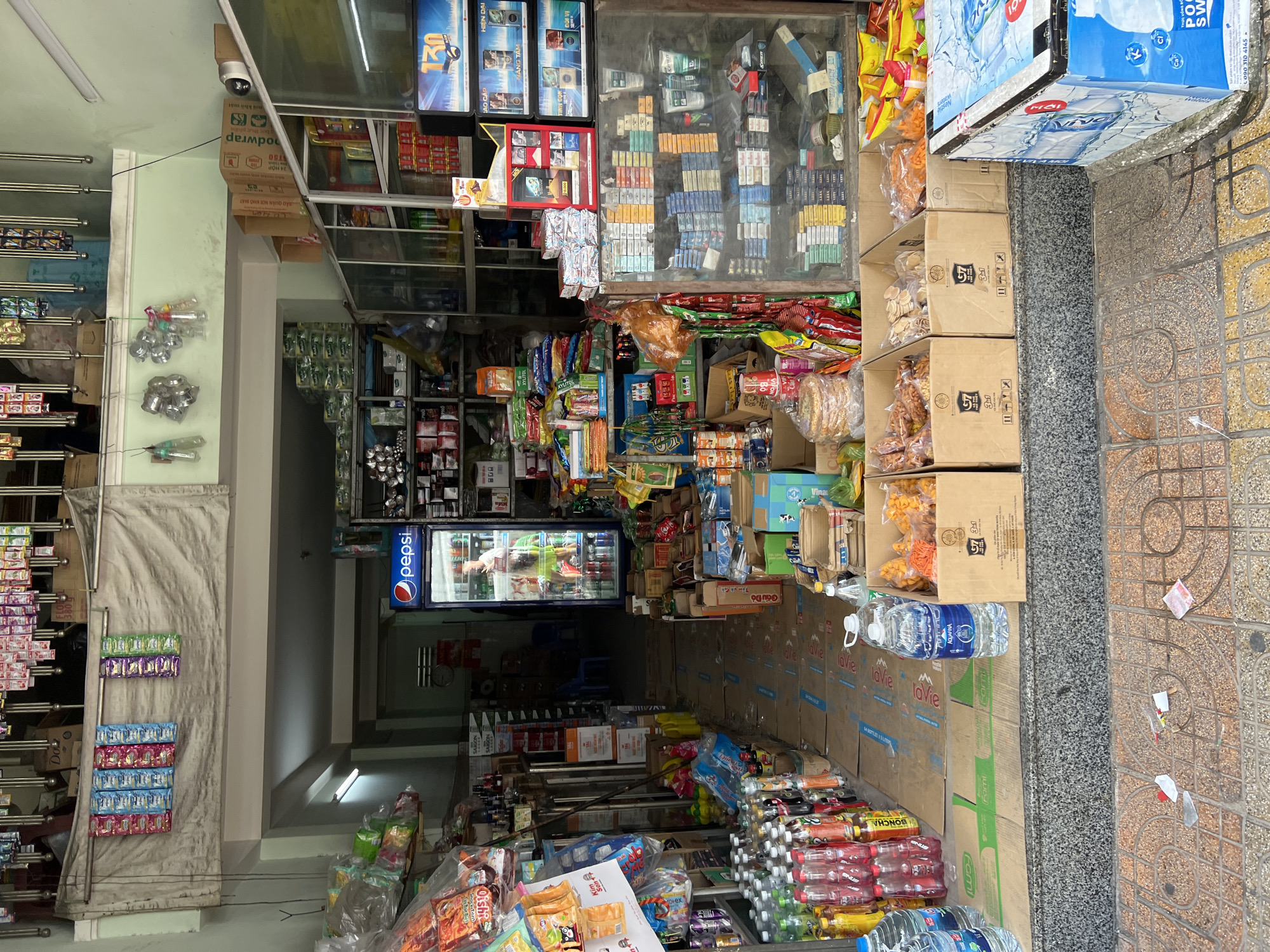 This image depicts the interior and entrance of a small convenience store in Nha Trang, Vietnam. The store appears to be well-stocked with a variety of packaged goods, including beverages, snacks, and cigarettes, arranged on shelves and in boxes. A Pepsi refrigerator is visible near the entrance, with a person standing inside it, seemingly stocking or retrieving items. The floor of the store is covered with cardboard, and the entrance opens to a sidewalk with people walking by. The overall scene suggests a typical street-level retail establishment in a bustling urban environment.