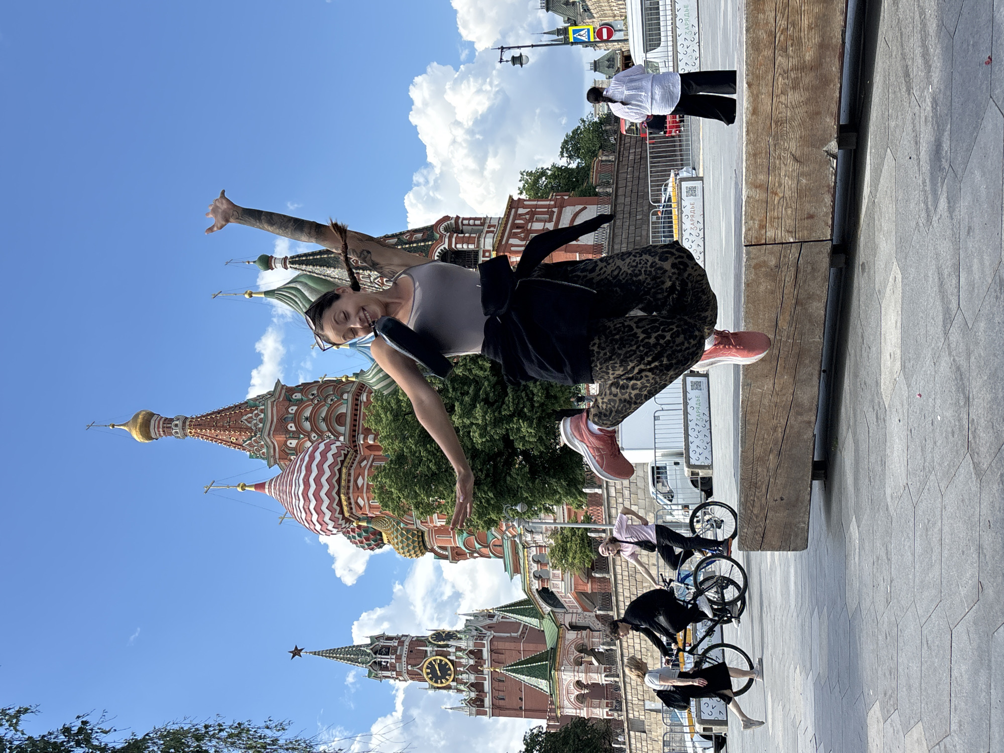 A woman with pigtails and a tattoo on her right arm is jumping in front of St. Basil's Cathedral and the Spasskaya Tower in Moscow, Russia. She is wearing a gray tank top, leopard print pants, and pink sneakers, and has a black jacket tied around her waist. Her right arm is raised. Several people on bicycles are visible to the right of the image, and a person in a white shirt and black pants is standing further back. The background includes blue skies with white clouds.