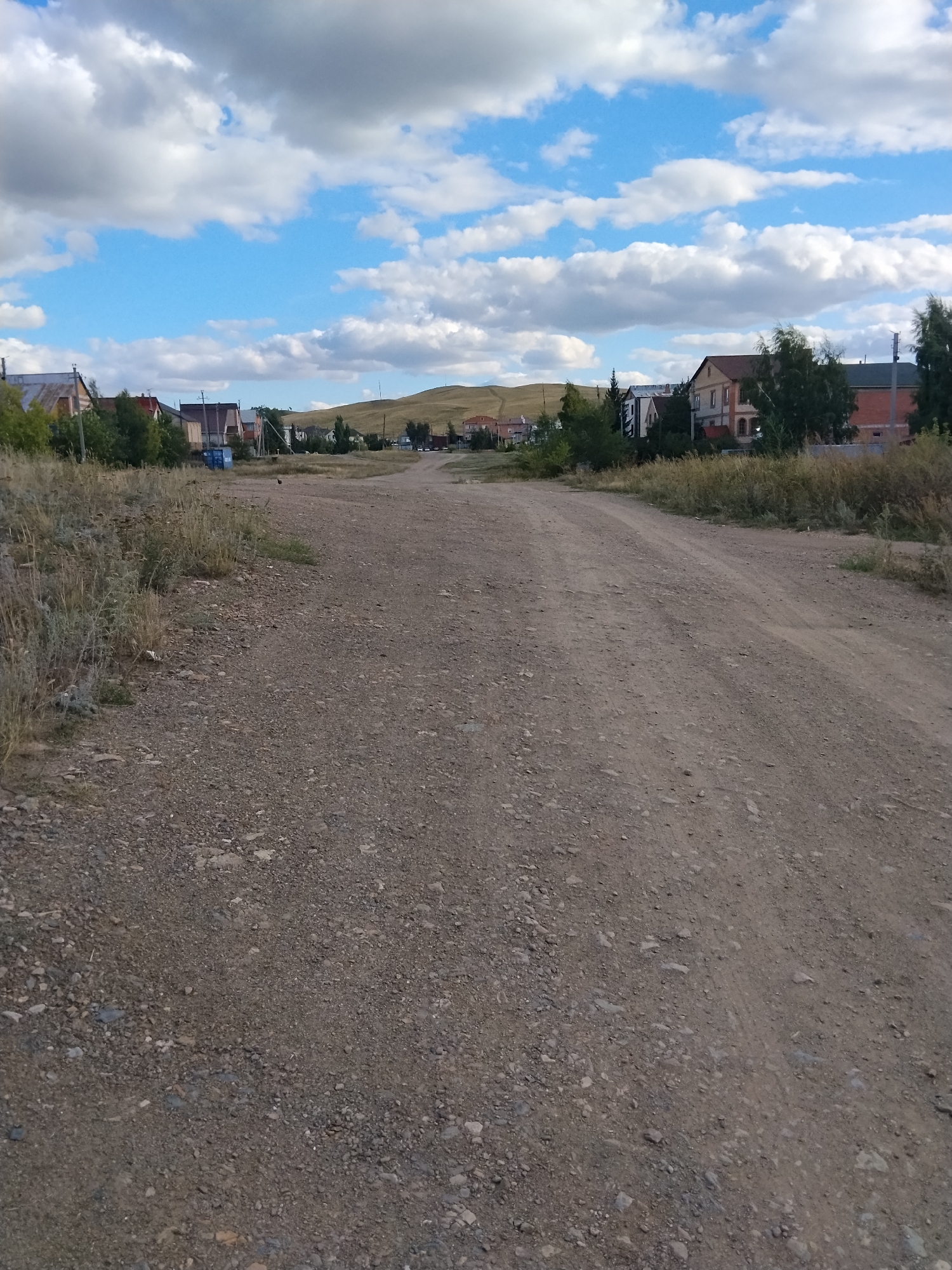 The image depicts a wide, unpaved dirt road stretching into the distance in a rural or suburban area of Kokshetau, Kazakhstan. The road is composed of gravel and dirt, with tire tracks visible. To the left of the road, dry, tall grass and weeds grow sparsely. On the right side of the road, there are houses with various roof types, some appearing to be under construction or recently built, interspersed with trees and vegetation. In the background, a gentle, grassy hill slopes upwards, topped with more houses. The sky is a bright blue, scattered with fluffy white clouds. The overall impression is a quiet, developing area on a clear day.