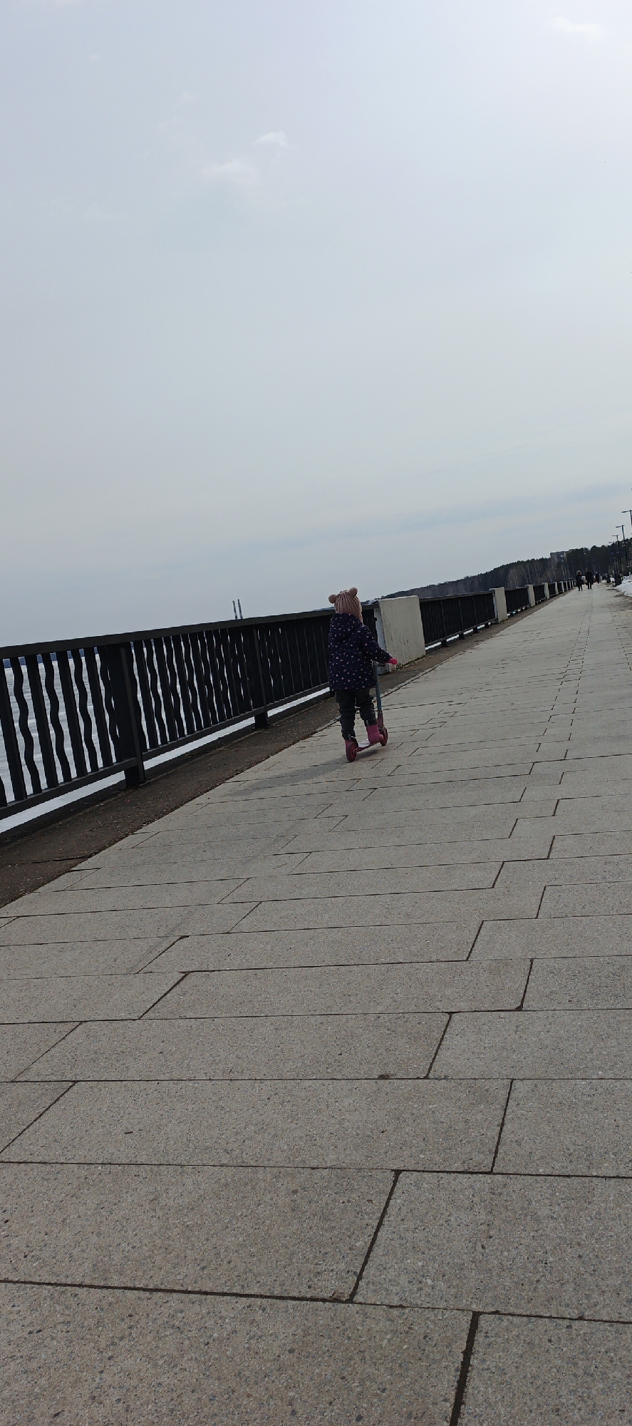 A child wearing a hat with ears, a blue patterned jacket, and pink boots is riding a scooter on a paved walkway. The walkway runs alongside a dark metal railing, with water visible beyond. In the distance, silhouettes of structures are faintly discernible. The location is Chaikovsky, Russia. The sky is overcast. Other people are walking along the path in the distance.
