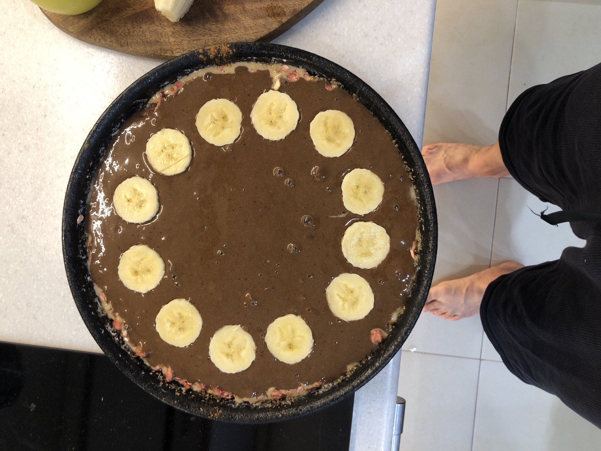 A high-angle, eye-level shot captures a person's feet and a circular pan containing what appears to be a chocolate pancake batter topped with sliced bananas. The pan is placed on a speckled white countertop, with a black stovetop visible to the left and a wooden cutting board with a sliced apple to the right. The pan's rim shows traces of what might be strawberries or other red fruit mixed with the batter. The person's bare feet and black pants are in the foreground, indicating they are standing in a kitchen. The scene is indoors. The image does not provide any specific information that would allow for the determination of its location within the city of Saint Petersburg, Russia.