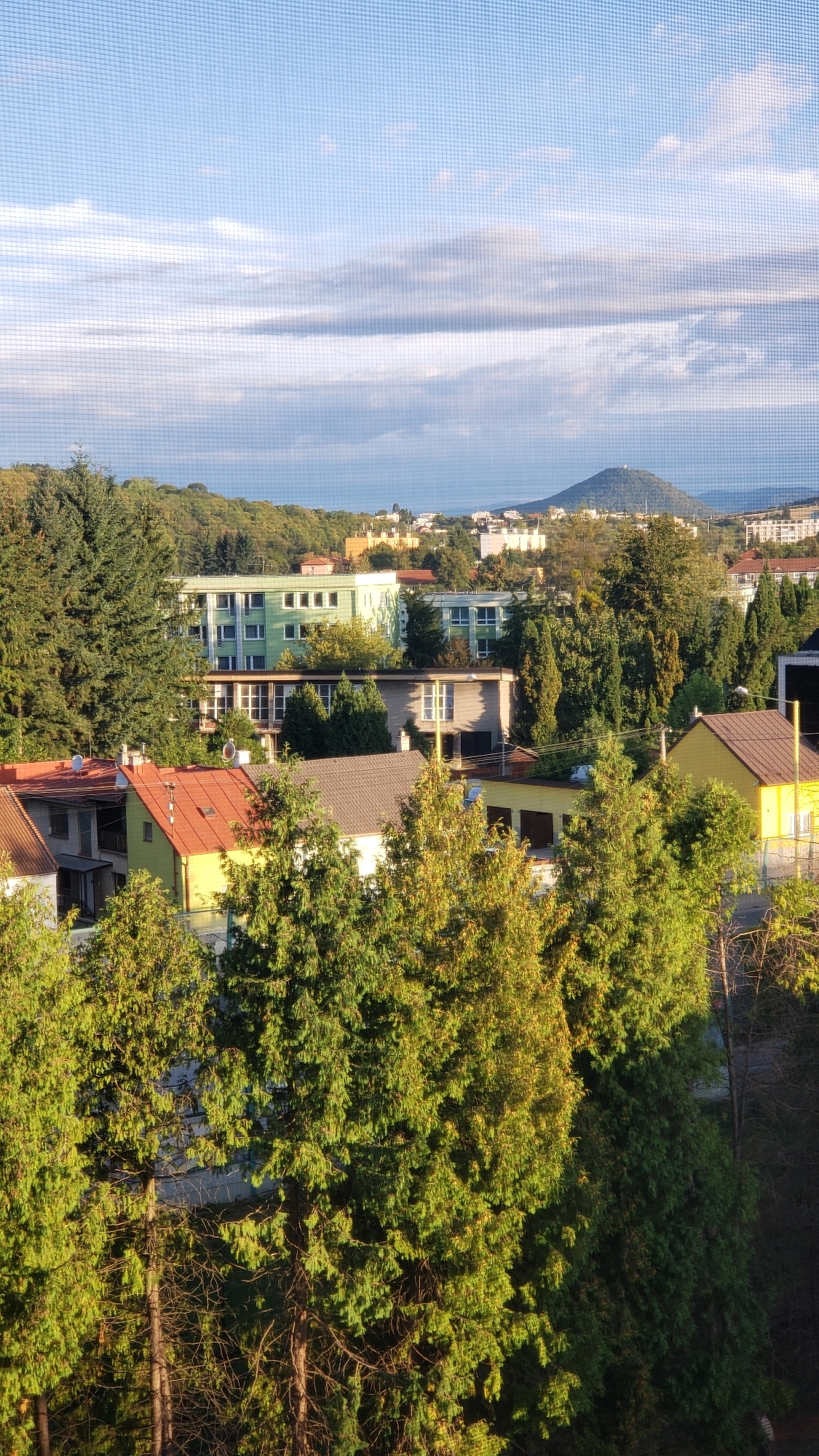 The image depicts a residential area in Prešov, Slovakia, with a view of a hill in the background. The foreground is dominated by lush green coniferous trees, some of which are in direct sunlight, casting shadows. Behind the trees, various residential buildings are visible, including a light green apartment building with multiple floors and windows, a row of smaller houses with red and brown roofs, and a yellow house. In the middle ground, there are more trees and buildings, including what appears to be a multi-story structure with a flat roof. The background features a distant, tree-covered hill with a structure on its summit, and the sky is partly cloudy with a blue hue. The scene is captured through a fine mesh screen, creating a slightly textured overlay on the image.