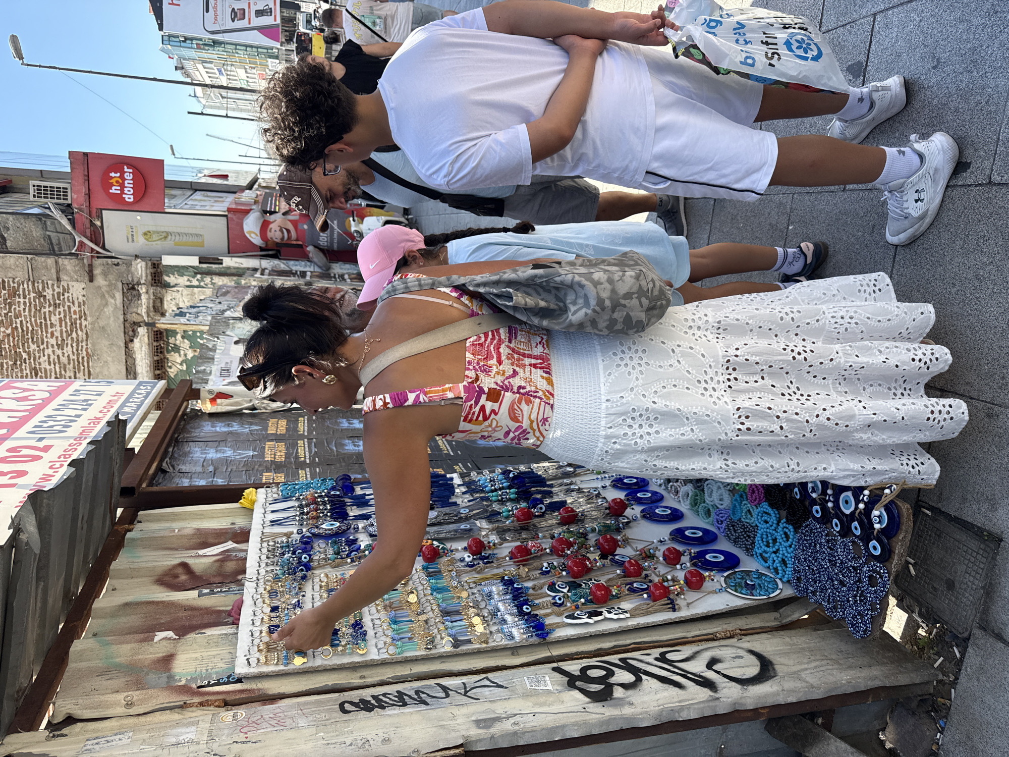 In Kadıköy, Turkey, a woman is examining a display of blue evil eye charms and other trinkets at a street market stall. She is wearing a white eyelet skirt and a floral top. Beside her, a young girl in a light blue t-shirt and pink baseball cap looks on, as does a man in a baseball cap and t-shirt. To the right, a young man in a white t-shirt and shorts is holding a plastic bag with Turkish writing. The stall is set up against a corrugated metal wall covered in graffiti. Behind the stall, a red sign for "hot döner" is visible. The scene takes place on a city sidewalk with pedestrians walking by.