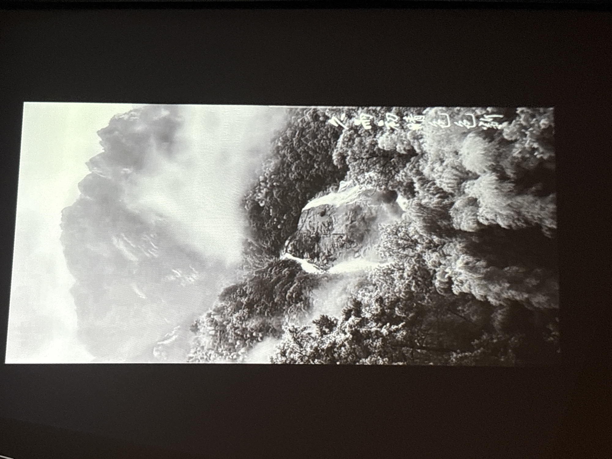 The image shows a black and white photograph of a misty mountain landscape. A waterfall cascades down a rocky slope amidst dense trees, some of which appear to be covered in snow or frost. The upper part of the image is dominated by rugged mountain peaks partially obscured by clouds and mist. To the right of the image, there is vertical text in Chinese characters. This photograph is a projection on a dark surface, indicating it's likely being displayed in a presentation or screening. The specific location in Pudong, China is not evident from the image itself.