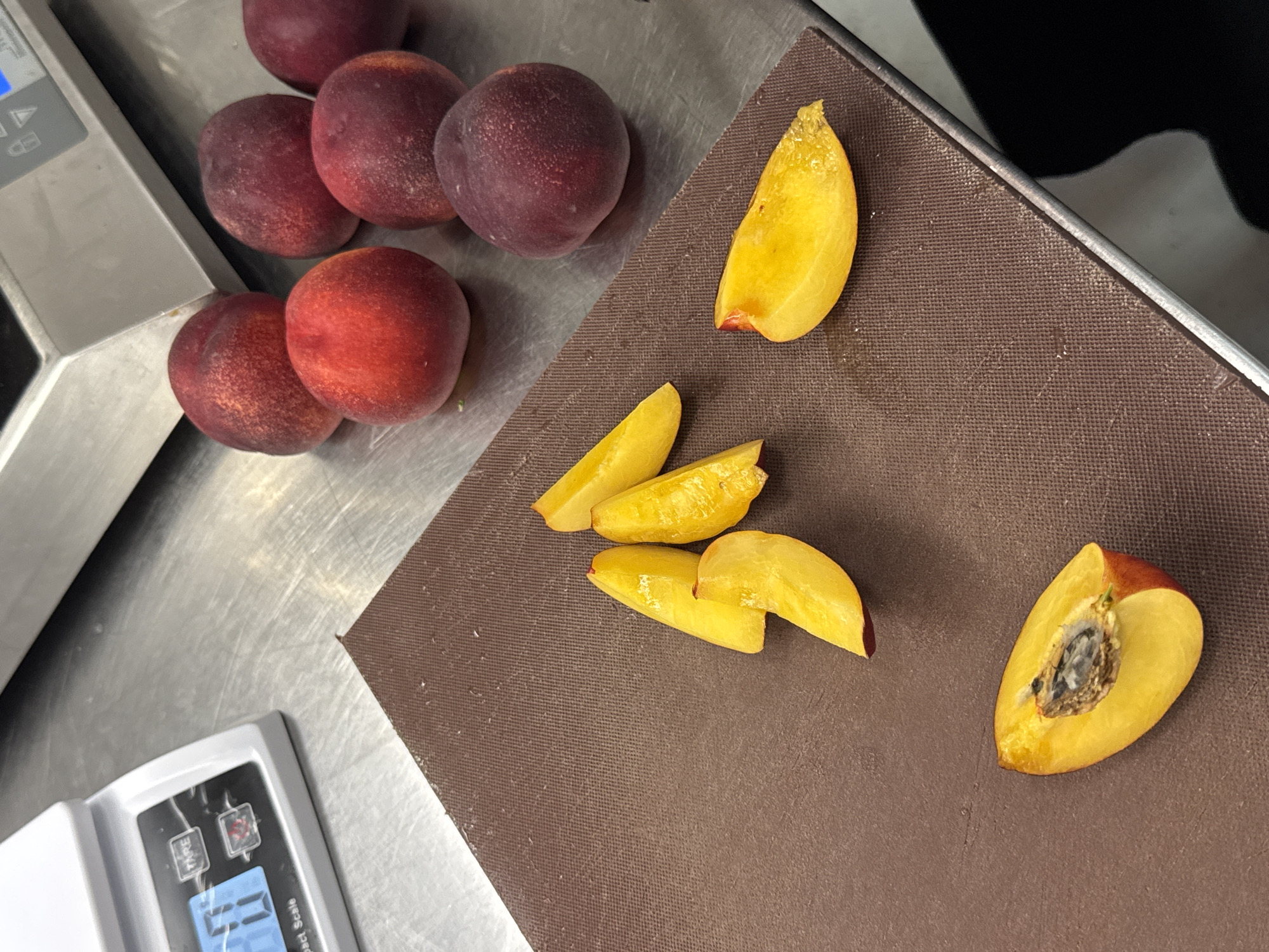 The image shows a close-up of several whole peaches and sliced peach wedges on a dark brown cutting board. The peaches are a mix of red and yellow, with some showing a deep burgundy hue. The sliced wedges are bright yellow and appear juicy, with one wedge revealing the peach's pit. To the left of the cutting board, a white digital scale with a blue display is visible. In the background, more whole peaches are scattered on a stainless steel surface. The lighting is bright, suggesting an indoor setting, possibly a kitchen. There are no people visible in the image. The image does not provide any information that would allow it to be placed in Belgrade, Serbia.