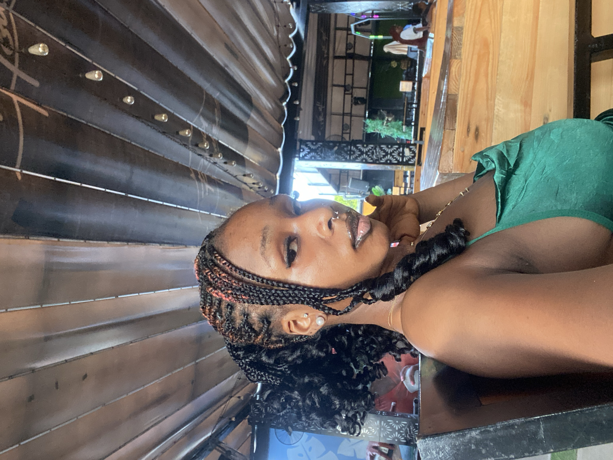 A medium shot captures a young Black woman from the chest up, seated at a table in what appears to be an outdoor, covered seating area. She is facing the camera and has dark skin, her hair styled in braided pigtails with some strands featuring pink highlights. She is wearing a bright green, sleeveless, ruched dress. The woman has on makeup, including eyeliner and lip gloss, and sports a nose ring and an earring in her left ear. Her right hand is raised, with her fingers touching her chin, as she gazes directly at the viewer.
The background shows a wooden ceiling with light fixtures. To the left, a blue wall with a white patterned design is visible. To the right, a rustic wooden bench and table setup is apparent, with other patrons and a stage area in the distance. The overall setting suggests a casual dining or entertainment establishment. The location in Howayi, Nigeria is not explicitly indicated by any visual cues in the image.