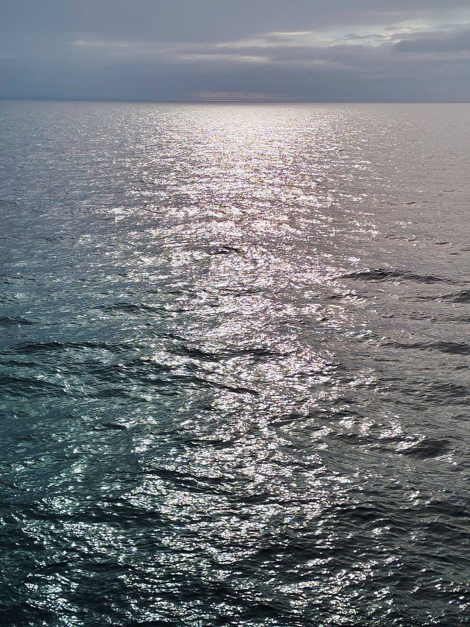 The image shows a body of water, possibly a sea or a large lake, with sunlight reflecting off the surface. The water's surface is slightly choppy, creating a sparkling effect. In the background, there is a visible horizon line separating the water from the sky, which appears cloudy. No specific information about the location in the city of Unknown, Russia, is discernable from the image. There are no people, subjects, or objects explicitly identifiable other than the water and the sky. There are no actions or interactions visible.