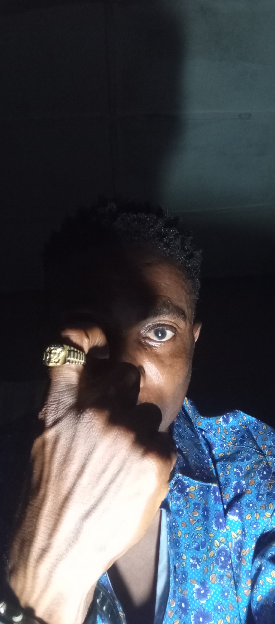 A close-up, low-light, self-portrait style image depicts a person with dark skin and curly hair. The individual is wearing a blue collared shirt with a floral pattern. A gold ring with an engraved face is visible on the person's right hand, which is held up near their face, obscuring part of it. The background is dark and indistinct. The overall lighting creates strong shadows, particularly on the face. The person appears to be looking directly at the camera. The image is believed to have been taken in Adankolo, Nigeria.