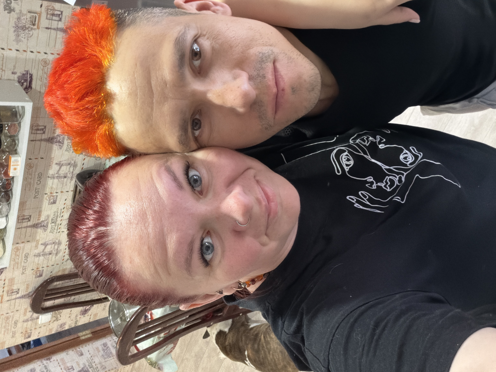 A selfie taken by a red-haired woman and an orange-haired man. The man and woman are lying down, with the man on top. The woman is wearing a black shirt with a white cartoon image on it. The man is wearing a black shirt. The background is a table with a glass container with spices and various objects behind. The bottom right corner of the image shows a dog resting under the chair. The location is undetermined; there is no indication it is Chaikovsky, Russia.
