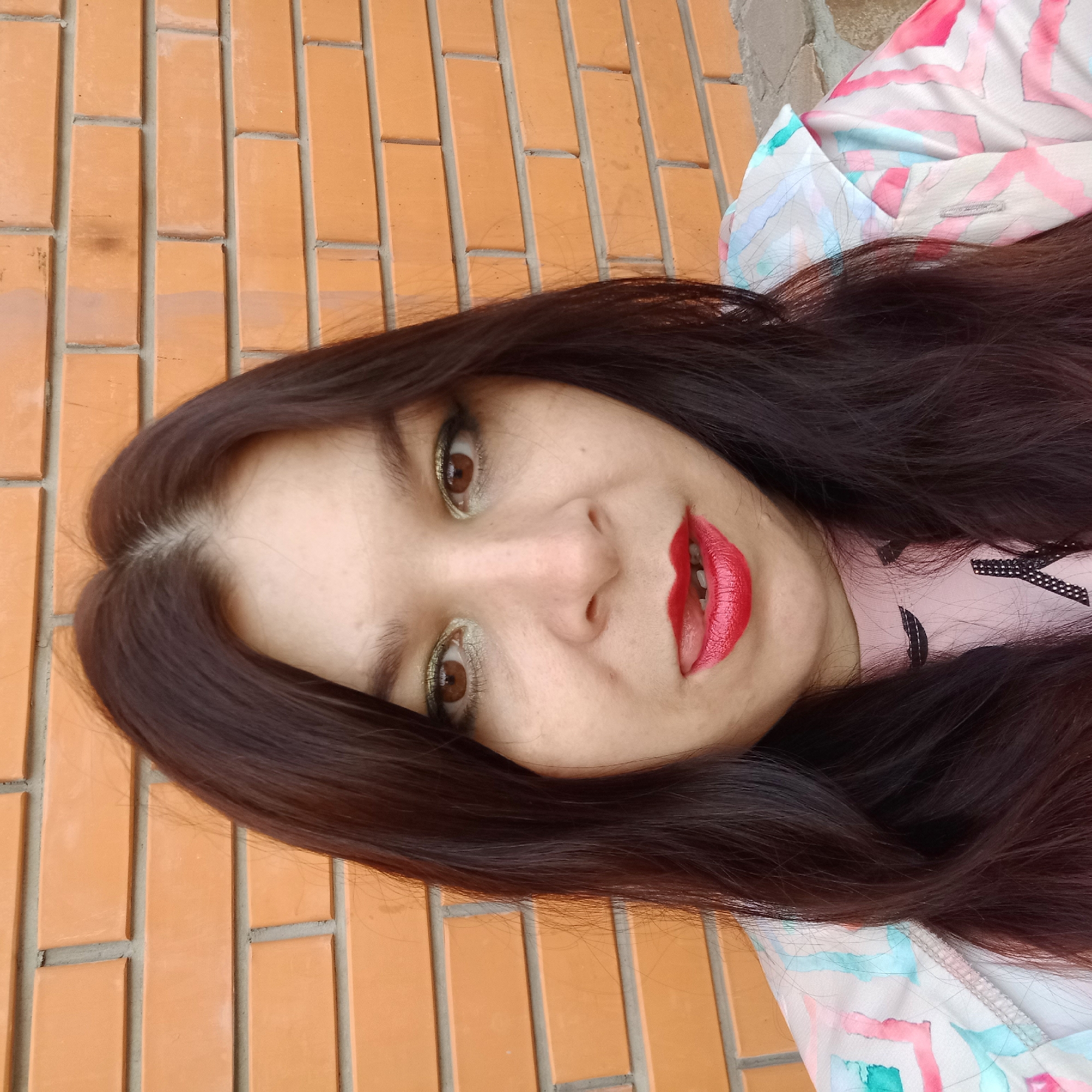 A woman with dark hair and brown eyes, wearing red lipstick, is pictured close-up against a brick wall. She is sticking her tongue out slightly. She is also wearing a pink shirt and a colorful jacket with geometric patterns. The scene is outdoors, with the brick wall forming the background. The location is the city of Kropyvnytskyi, Ukraine.