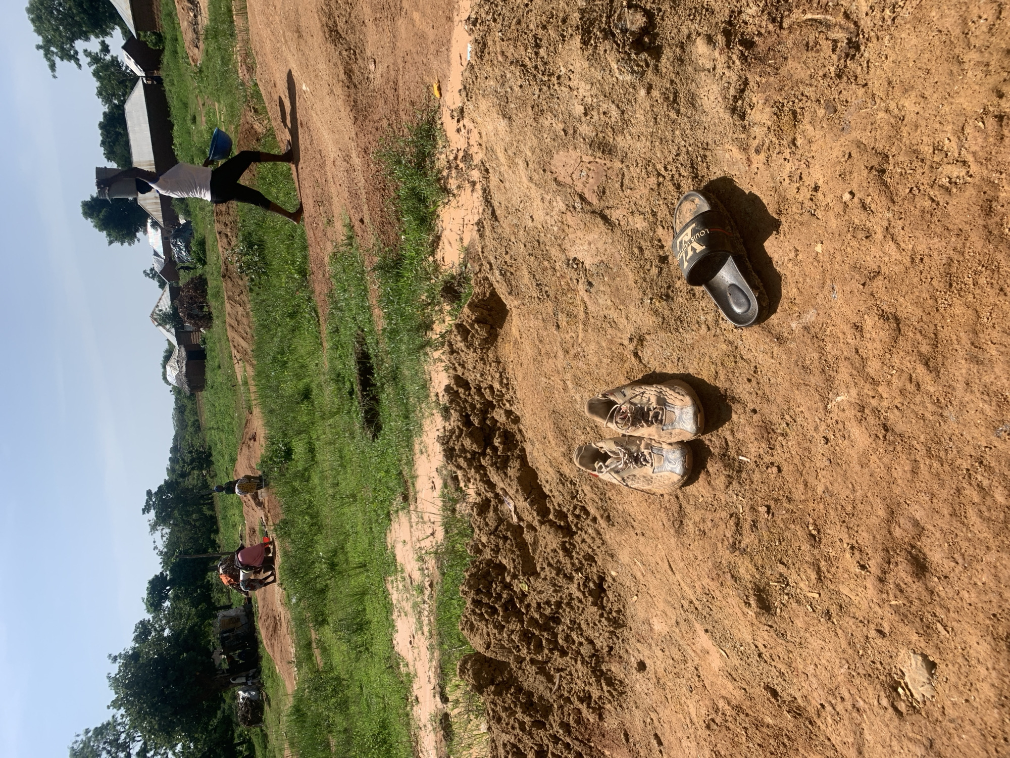 The image depicts a sun-drenched outdoor scene in Buga da Gindi, Nigeria. In the foreground, a pair of worn, light-colored athletic shoes and a black slide sandal are resting on a mound of reddish-brown soil. The ground is a mixture of loose dirt and some dry vegetation. In the middle ground, several people are engaged in activity, some appearing to be working with sand or soil. One woman is visible in the background, carrying a bucket on her head and a blue container in her hand, walking away from the viewer. The environment consists of sparse vegetation, with trees and bushes scattered across the landscape, and a collection of simple structures with corrugated metal roofs in the distance, suggesting a rural or semi-rural settlement. The sky is clear and blue, indicating fair weather.