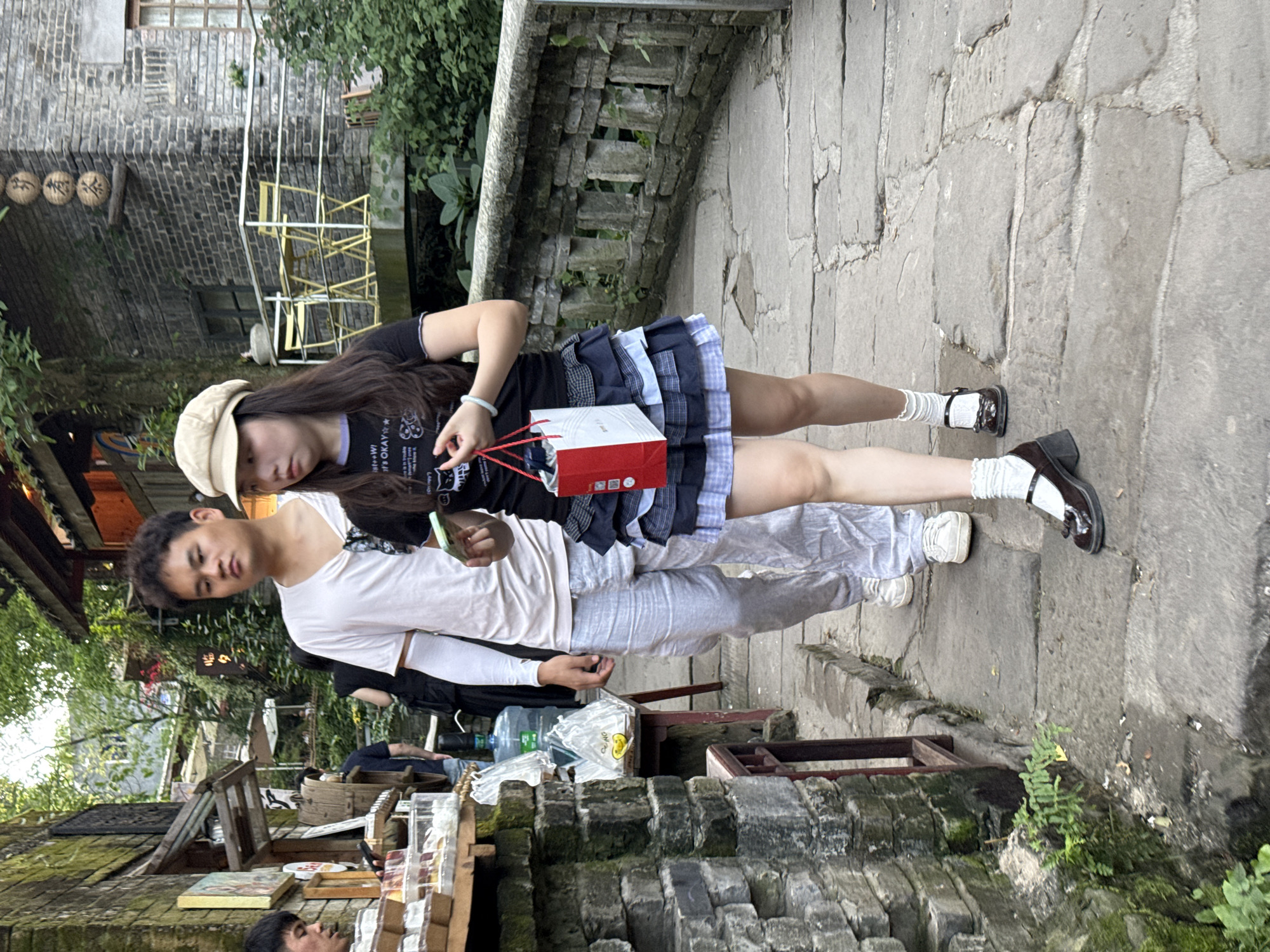 In Beibei, China, two young people stand on a stone path. The woman wears a black t-shirt, a short, layered skirt with shades of blue, white socks, and black Mary Jane shoes. She holds a red and white shopping bag. On her head is a tan baseball cap. The man wears a white t-shirt and light grey pants. They appear to be standing near an outdoor market or vendor area with brick structures and various goods on display. Greenery surrounds the area.