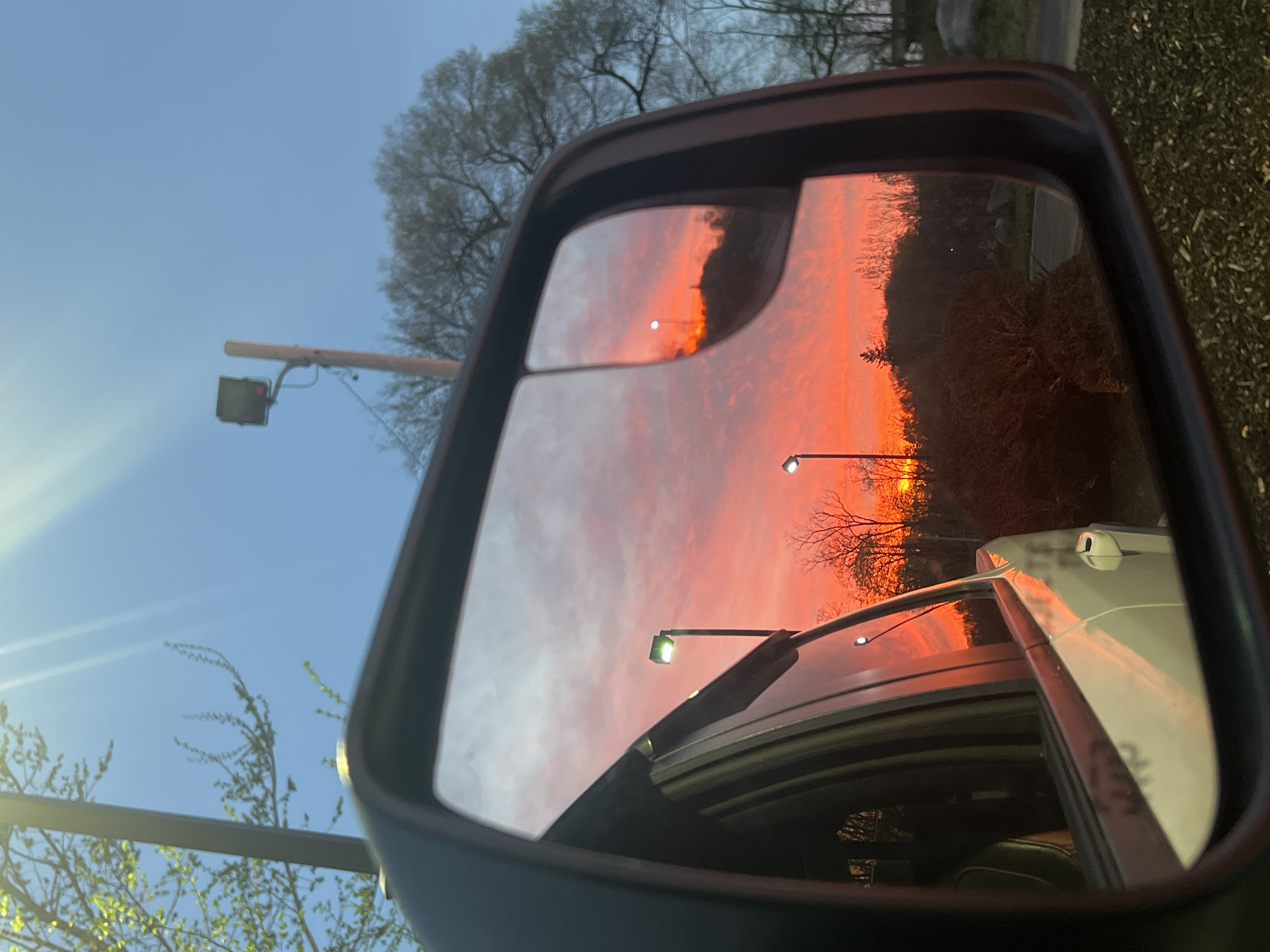 The image depicts a side mirror of a vehicle, reflecting a sunset scene. The reflection showcases a vibrant orange and red sky, indicating either sunrise or sunset. There are trees and lampposts visible in the background of the reflection. The foreground of the image contains elements outside the reflection, including trees and the surrounding environment. The photo was taken somewhere in the city of Newburgh, United States of America.