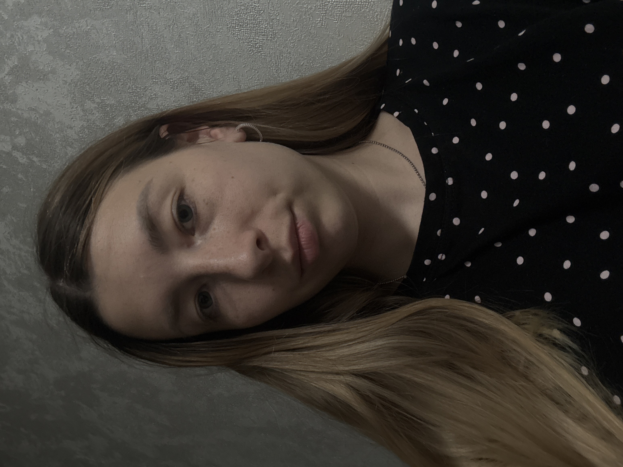 A woman with long, blonde hair is lying down against a gray wall. She is wearing a black shirt with white polka dots. There is a silver earring in her ear and a thin silver necklace around her neck. The image appears to be taken indoors. There is no information to suggest the location is in Moscow, Russia.