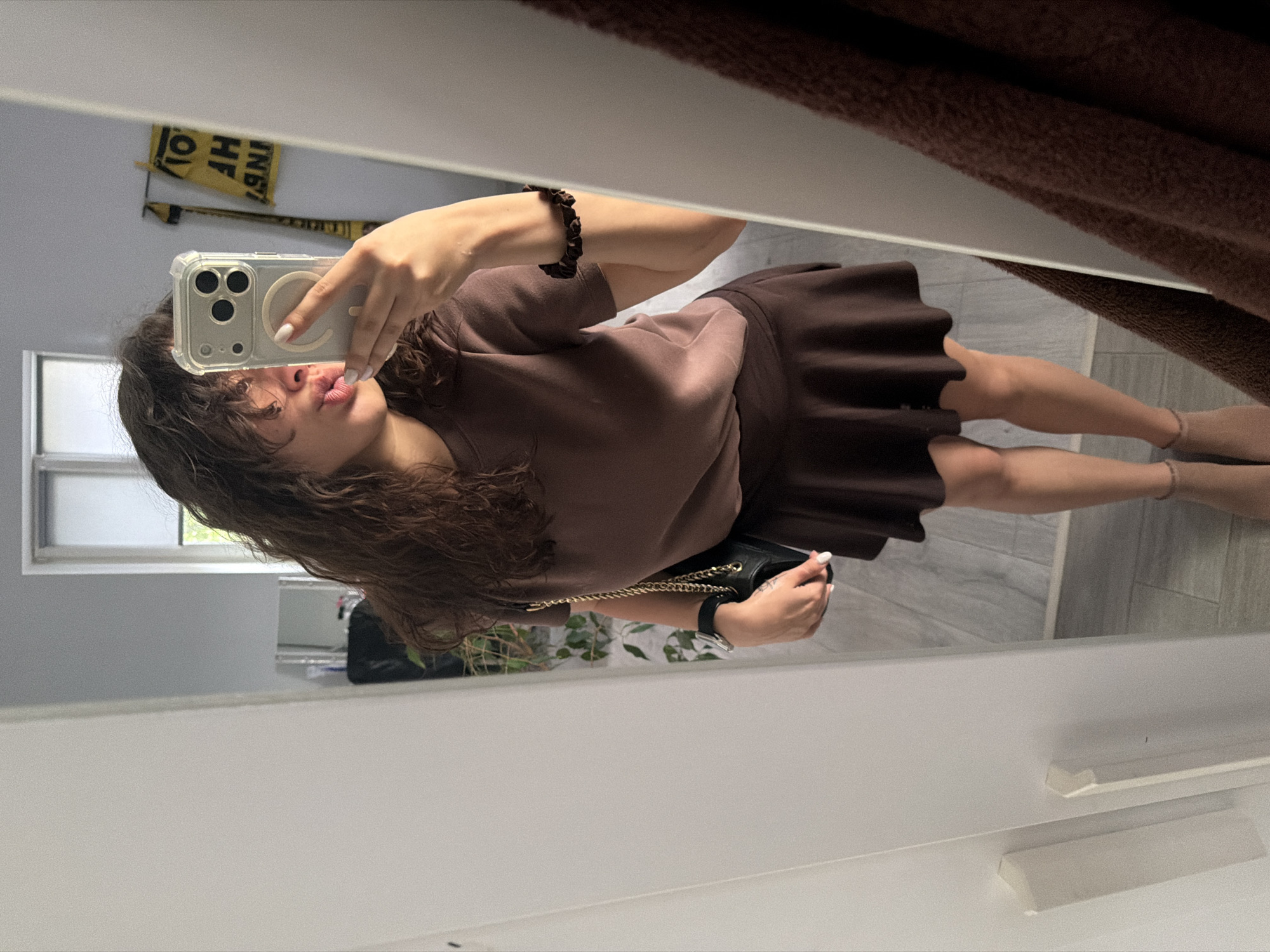 A young woman with long, curly brown hair is taking a mirror selfie indoors. She is wearing a brown, short-sleeved t-shirt and a matching brown pleated skirt. She is holding a clear phone case with a silver phone in her right hand, angled to capture her reflection. She has her lips puckered for a kiss. On her left wrist, she wears a black watch. A black purse with a gold chain strap is slung over her left shoulder and held by her left hand. She has on tan ankle socks and tan shoes. In the background, a window with white blinds is visible, as well as a yellow sign with black lettering. To the right of the woman, a brown towel hangs on a mirror frame. The floor is tiled with a gray and white wood-grain pattern.
