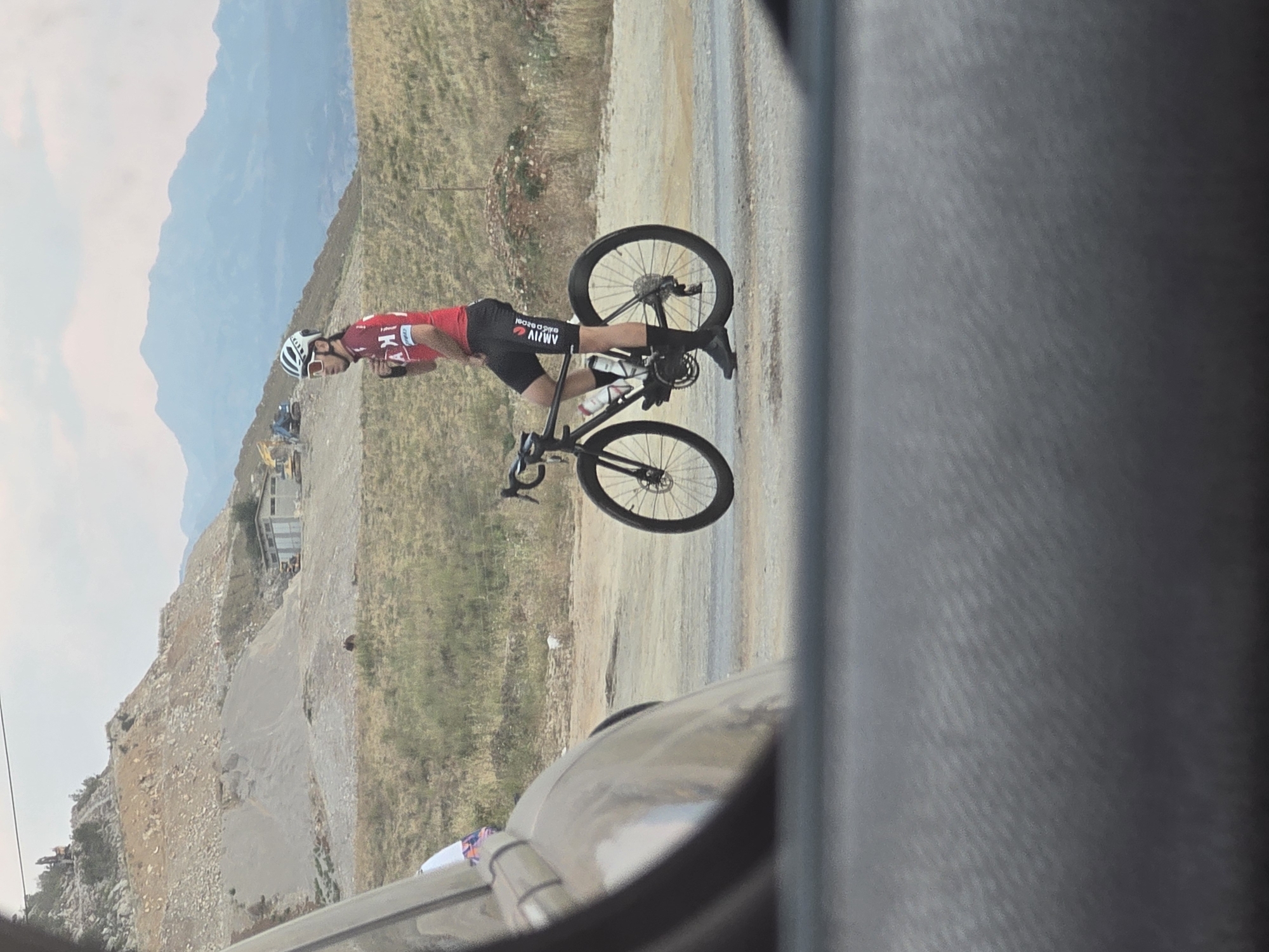 A cyclist wearing a red jersey and black shorts is standing next to his black road bicycle on a dirt road. He is wearing a white helmet and sunglasses, and is looking to his left. The bicycle has two water bottles in its cages. In the background, there is a quarry with heavy machinery and a building. Mountains are visible in the far distance. The scene appears to be on the outskirts of Orljevo, Montenegro, with a view of the rugged terrain and construction activity.