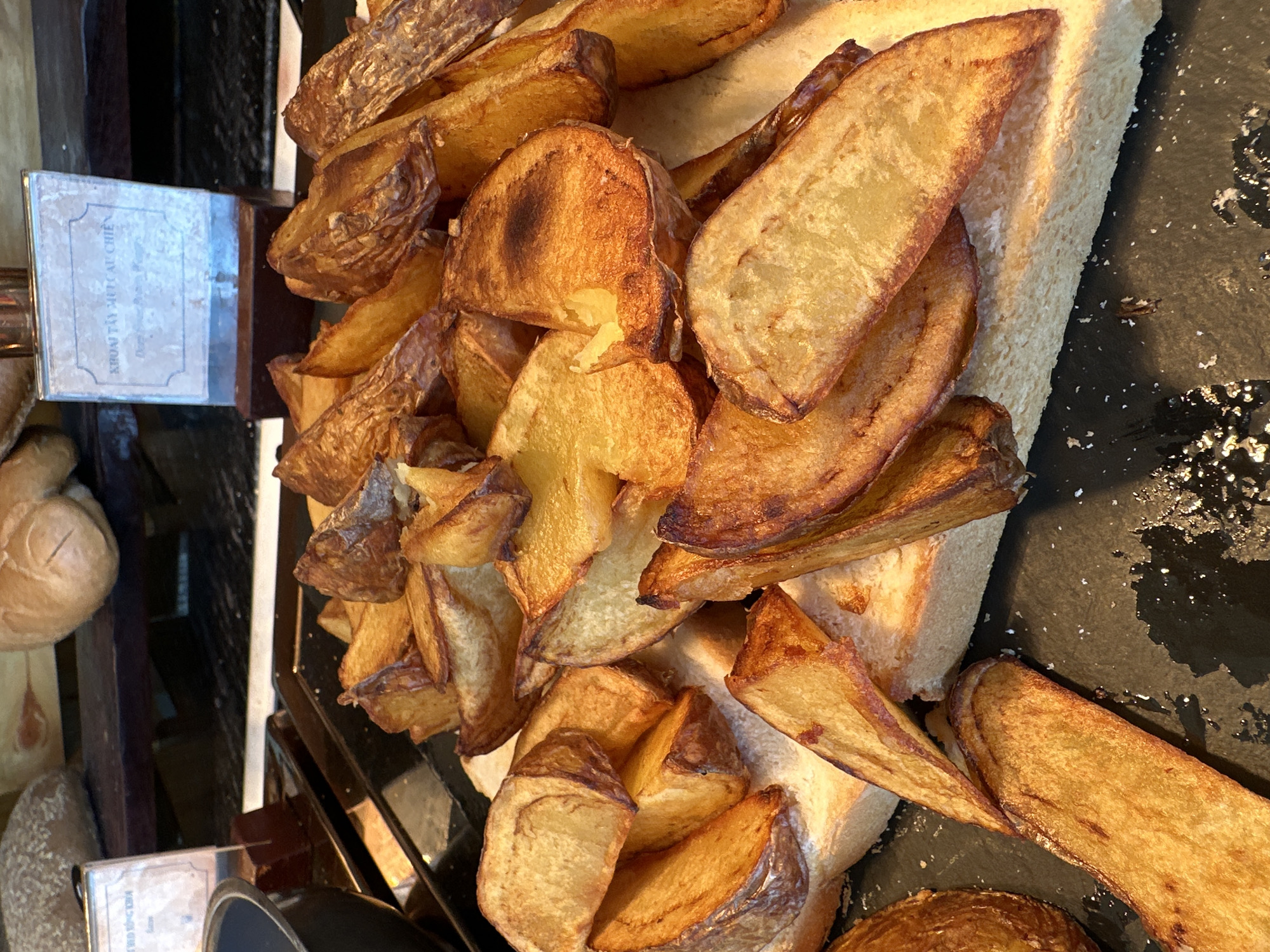 A detailed view captures a self-service food station featuring deep-fried potato wedges at a buffet in Nha Trang Ward, Vietnam. The primary subject is a substantial pile of golden-brown potato wedges, many with their skins intact and exhibiting crispy, dark brown edges. These wedges are arranged on top of several slices of plain white bread, which serve as an absorbent base on a rectangular black serving tray. Scattered white crystalline particles, consistent with salt, are visible on the black surface, particularly in the lower right quadrant.
Two informational signs with silver frames and dark brown bases are positioned behind the food. The sign on the right displays Vietnamese text: "KHOAI TÂY MÚT CÂU CHIÊN," followed by partially visible English text below it. A second, similar sign is partially visible on the left side of the frame, showing only a portion of its Vietnamese text.
In the upper background, a wooden shelf holds various baked goods. A prominent round loaf of bread with a textured crust is visible on the left, alongside other lighter-colored bread items. The scene is illuminated by overhead lighting, highlighting the texture and color of the food items. No individuals are present in the image.