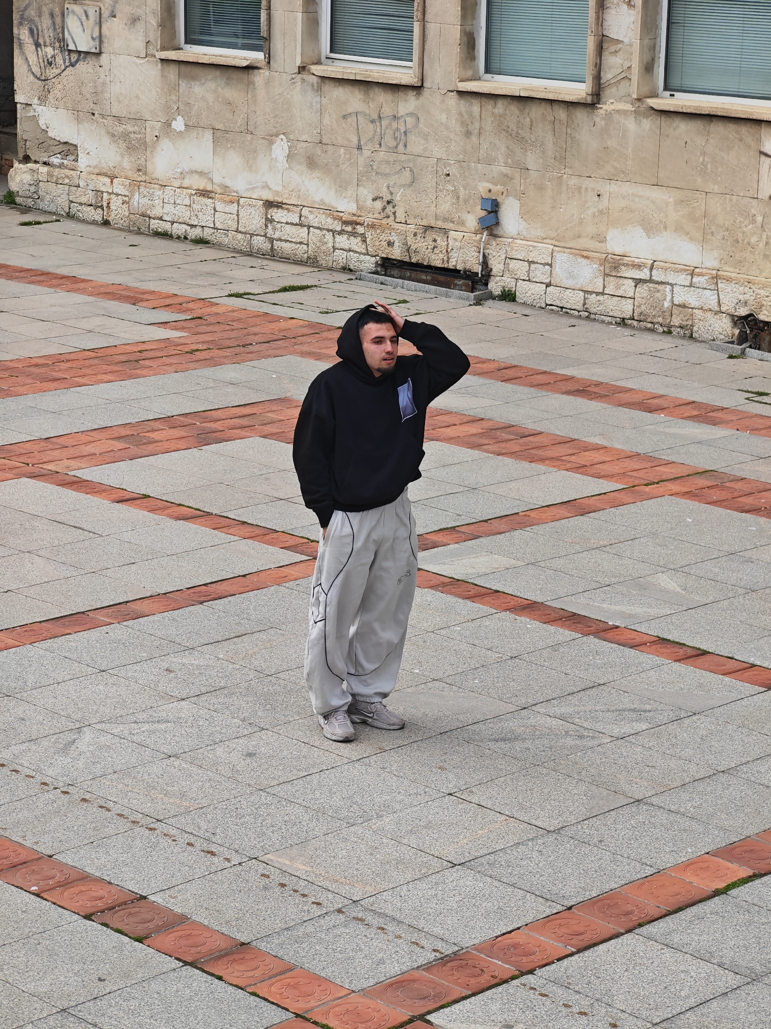 A young adult male stands centrally in an urban public square in Shumen, Bulgaria, the precise street or square name of which is not discernible from the image. He is wearing a black hooded sweatshirt with the hood up, light grey baggy sweatpants with black piping, and grey athletic shoes. A blue and white patch is visible on the left chest of his sweatshirt. His left hand is raised to the back of his head, touching his hair, and his mouth is slightly ajar. He gazes generally downward and slightly left from his perspective. The ground consists of light grey square tiles interspersed with diagonal lines of reddish-brown decorative embossed tiles. Small tufts of grass protrude between some tiles. The background features a two-story building facade with significant wear, exposed stone blocks at the base, and areas of damaged plaster revealing underlying material. Multiple instances of black graffiti are present on the upper wall. Two windows with white frames and drawn horizontal blinds are visible. A blue rectangular electrical fixture is mounted on the wall below the right window. The scene is illuminated by diffused light, suggesting an overcast sky. No other individuals or significant objects are present in the immediate vicinity.