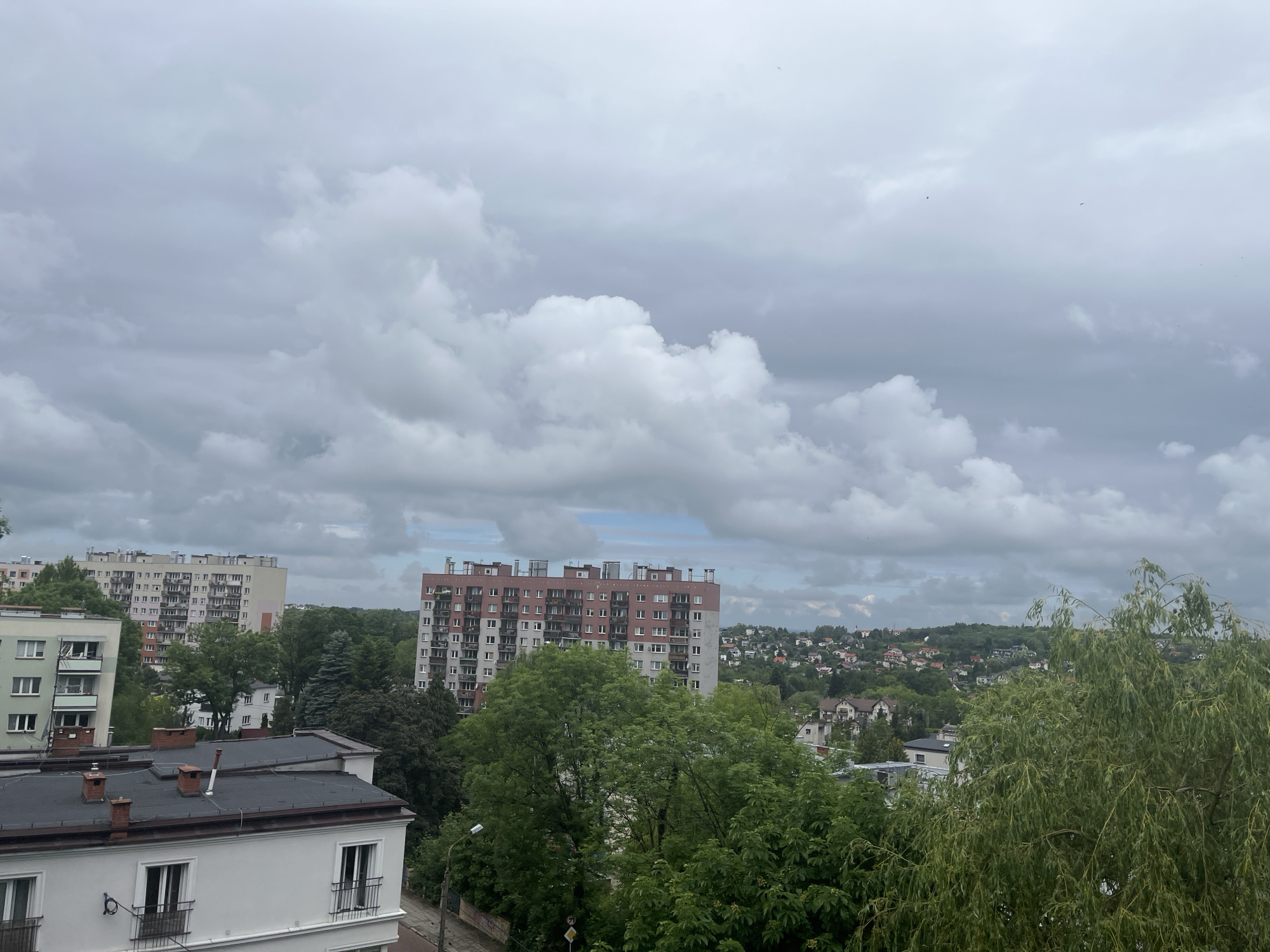 The image presents an elevated view of an urban or suburban landscape under a heavily overcast sky. The scene is dominated by residential buildings and dense greenery.
In the foreground, the top of a white building with a flat, dark roof and several brick chimneys is visible on the lower left, featuring windows with dark railings. In the mid-ground, two large, multi-story apartment blocks stand out. On the left, a light-colored building with many windows and balconies is partially visible. Towards the center-right, a taller apartment building with a reddish-brown upper section and a light grey facade is prominent, rising above the trees.
Abundant vegetation is scattered throughout the scene. A large, leafy tree with drooping branches, possibly a willow, occupies the right side of the frame. Various other dark green trees surround the mid-ground buildings. In the distant background, a hillside is covered with numerous smaller houses, characterized by reddish roofs, nestled amongst trees.
The sky is entirely overcast with extensive grey and white clouds, suggesting a cloudy or gloomy daytime. There is no direct sunlight, and the lighting is diffuse. A few tiny, dark specks, possibly birds, are visible in the upper sky. No people or visible text are present. The image depicts a built-up area integrated with significant natural elements.