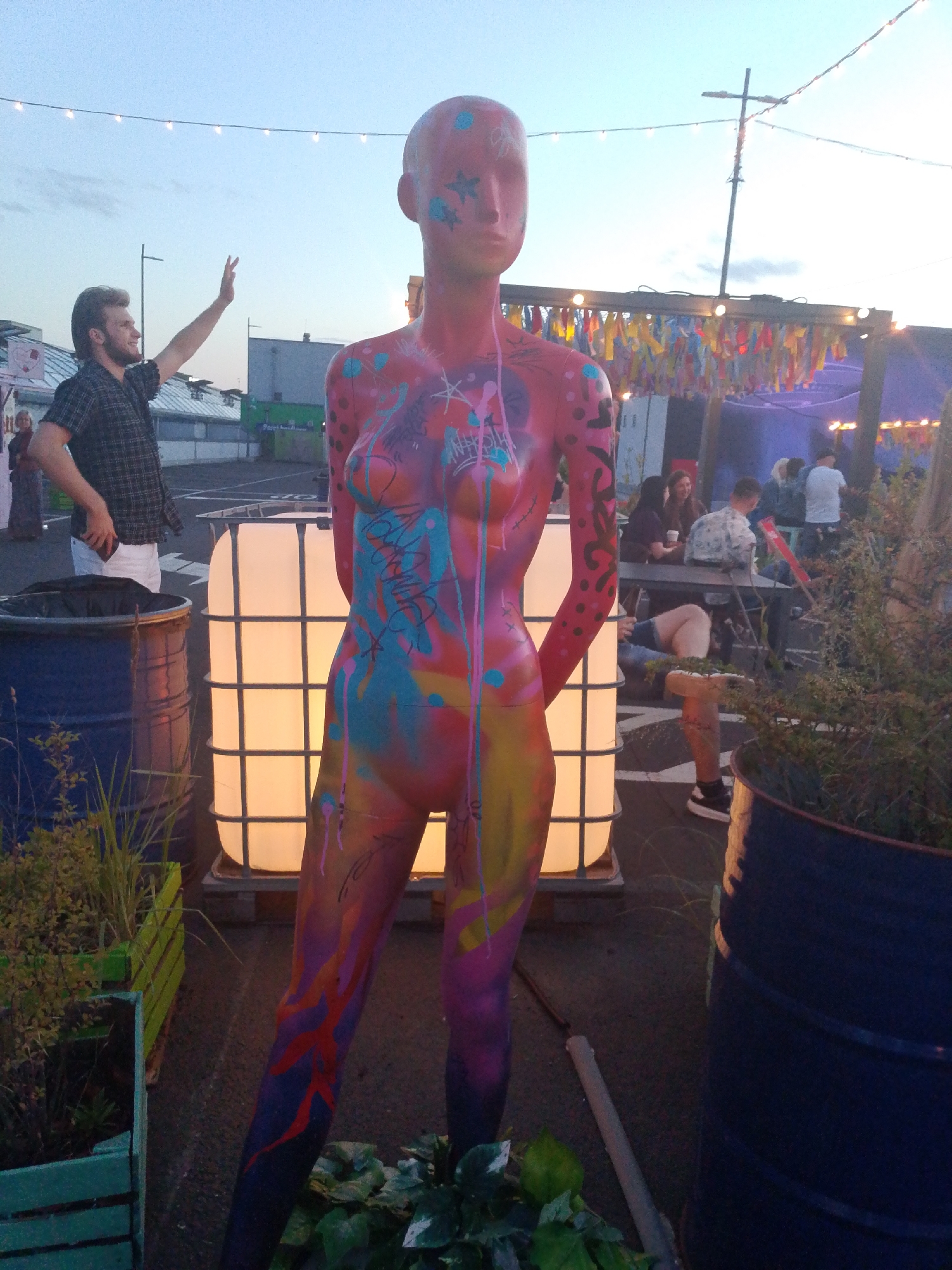 The image depicts an outdoor evening scene, possibly at a festival or gathering, with a stylized, brightly painted mannequin as the central subject. The mannequin is covered in vibrant graffiti-like art in shades of pink, blue, yellow, and orange, with some abstract patterns and what appear to be star shapes on its head and body. It stands in the foreground, facing forward, with its arms slightly bent and hands clasped at its waist.
Behind the mannequin, several objects are visible. To the left, a large blue barrel is partially obscured. Next to it is a tall, illuminated white translucent tank, possibly an IBC tote, emitting a warm glow. In the middle ground, a group of people are seated around tables under a structure decorated with colorful streamers and string lights, suggesting a social or entertainment event. The lighting indicates it is dusk or early evening, with string lights illuminated and the sky showing a soft twilight glow.
To the far left of the image, a young man with a beard wearing a plaid shirt and white trousers is captured mid-gesture, with his right arm raised and hand open, as if waving or signaling. His expression appears cheerful. Further in the background, buildings and structures are visible, but their details are less clear due to the distance and lighting. The ground appears to be paved. The overall atmosphere seems relaxed and festive, characteristic of an outdoor event in the evening. The location context is Wrocław, Poland. There is no visible text in the image.