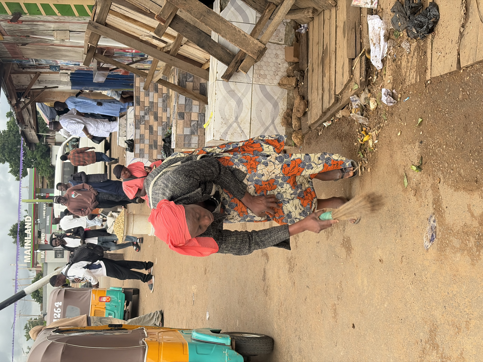 In the city of Gura Top, Nigeria, a woman wearing a red headscarf and a patterned orange and white dress sweeps a dusty, unpaved road with a straw broom. She is leaning forward, her face obscured by the movement of her work. A turquoise and yellow auto-rickshaw is parked to her left. Other individuals are visible in the background, including men in casual clothing and a gas station with "MAINLAND" signage. The scene appears to be an outdoor market or roadside area, with wooden structures, debris, and various goods for sale. The ground is littered with trash, including plastic bags and wrappers.