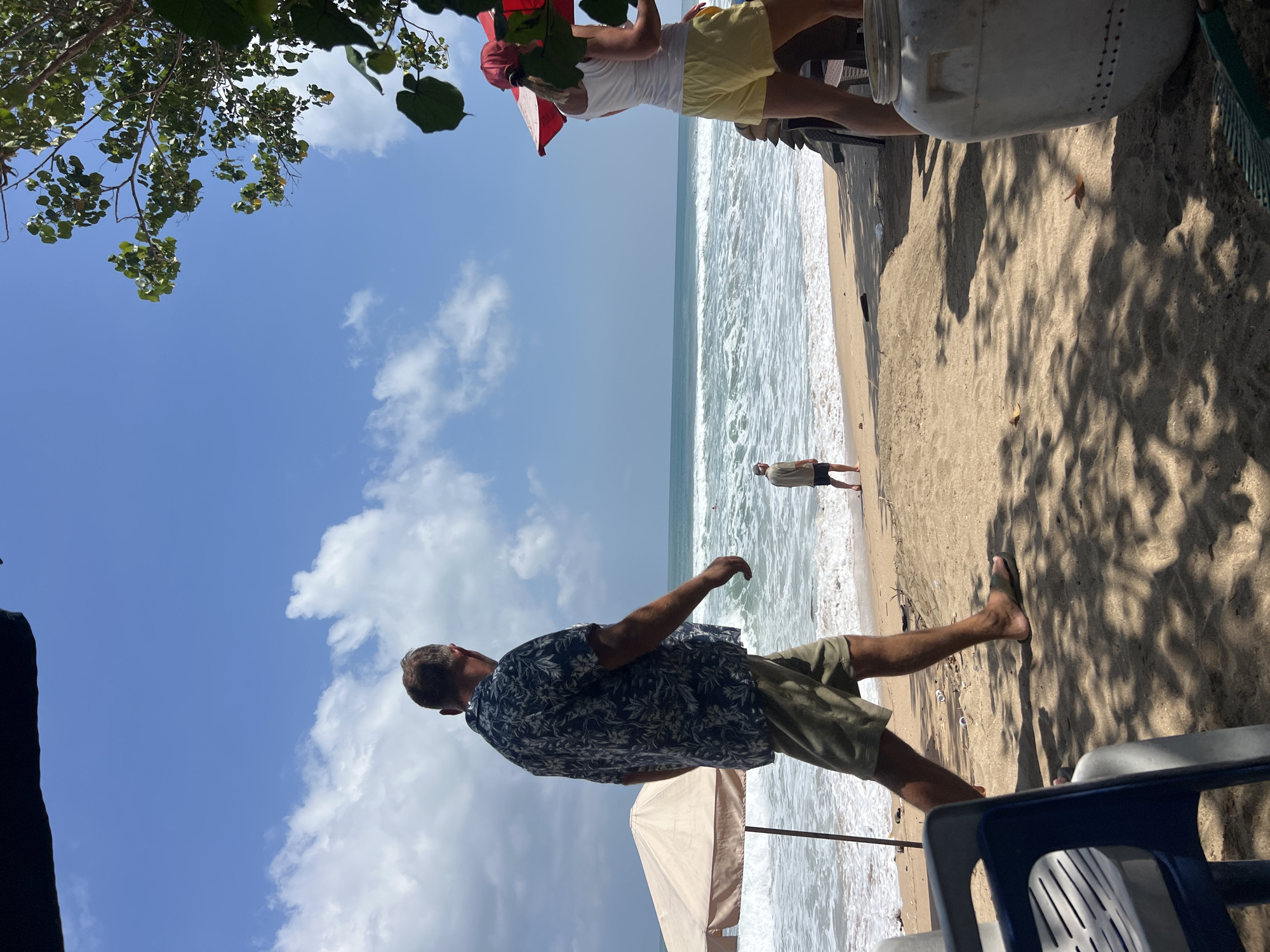 An outdoor medium shot shows a beach scene in Kuta, Indonesia, on a clear, sunny day with some scattered clouds. The ocean with moderate waves is visible in the background. In the foreground, a man wearing a dark floral shirt and khaki shorts walks to the left. To the right, a person wearing a white tank top and yellow shorts stands near a red umbrella. Further back on the beach, a man in a light-colored shirt and dark shorts stands facing the ocean. Beach chairs and a large white container are also present. The sand is light brown, and tree branches with green leaves cast shadows across the sand.