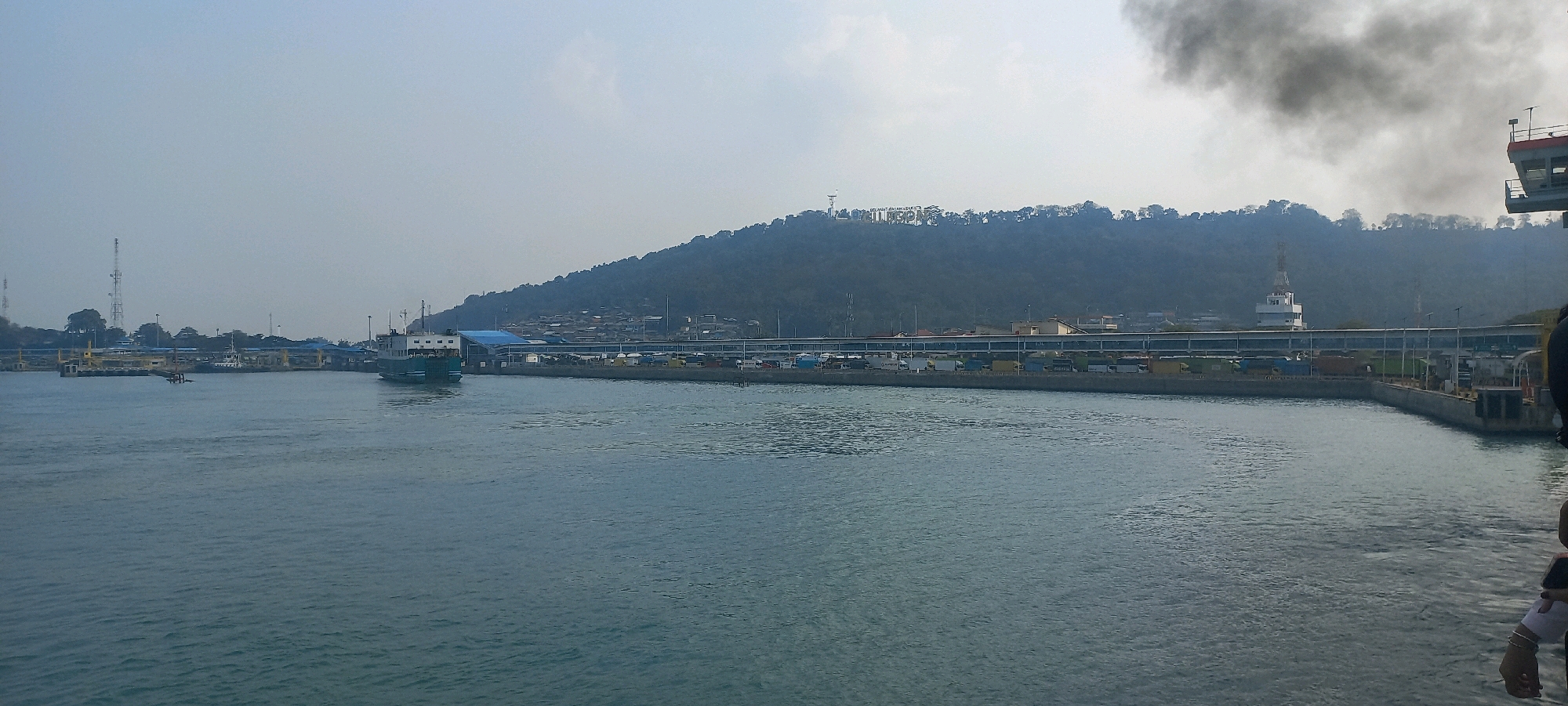 This is an outdoor, wide-angle panoramic photograph taken from a body of water, likely a harbor, looking towards a coastal area. The sky is overcast and hazy, with a plume of dark smoke rising from the right side of the frame, obscuring the upper part of a distant hillside.
In the foreground, the water is a deep blue-green with visible ripples. Along the middle ground, a port facility is visible. Several boats and ferries are docked or in the water, including a large green ferry on the left side of the frame. A long pier extends across the frame, lined with various vehicles, including trucks and what appear to be shipping containers. Buildings with blue roofs are situated behind the pier.
Dominating the background is a large, tree-covered hill. On the crest of the hill, a large, stylized sign spelling out "SELAMAT HARI KEMERDEKAAN" (Happy Independence Day) is visible in bright letters. Various structures, including what appears to be a tall tower or communication antenna, are scattered across the hillside. The city of Cilegon, Indonesia, is known for its industrial port and hilly coastal landscape, making this scene consistent with a view of its harbor and surrounding terrain. A portion of a ship or structure is visible on the far right edge of the image, and a person's arm and hand are partially visible in the bottom right corner.