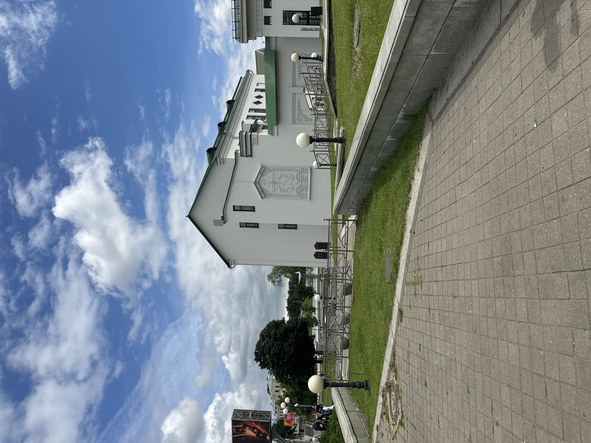 The image depicts a view of the Upper Town (Verkhniy Gorod) in Minsk, Belarus. The scene captures a white building with a green roof, featuring decorative architectural elements. In the foreground, a paved walkway leads towards a grassy area with lampposts and metal railings. A large billboard advertising a cultural event is visible on the left side of the frame, with several individuals walking in the vicinity. The sky above is blue with scattered white clouds, suggesting fair weather.