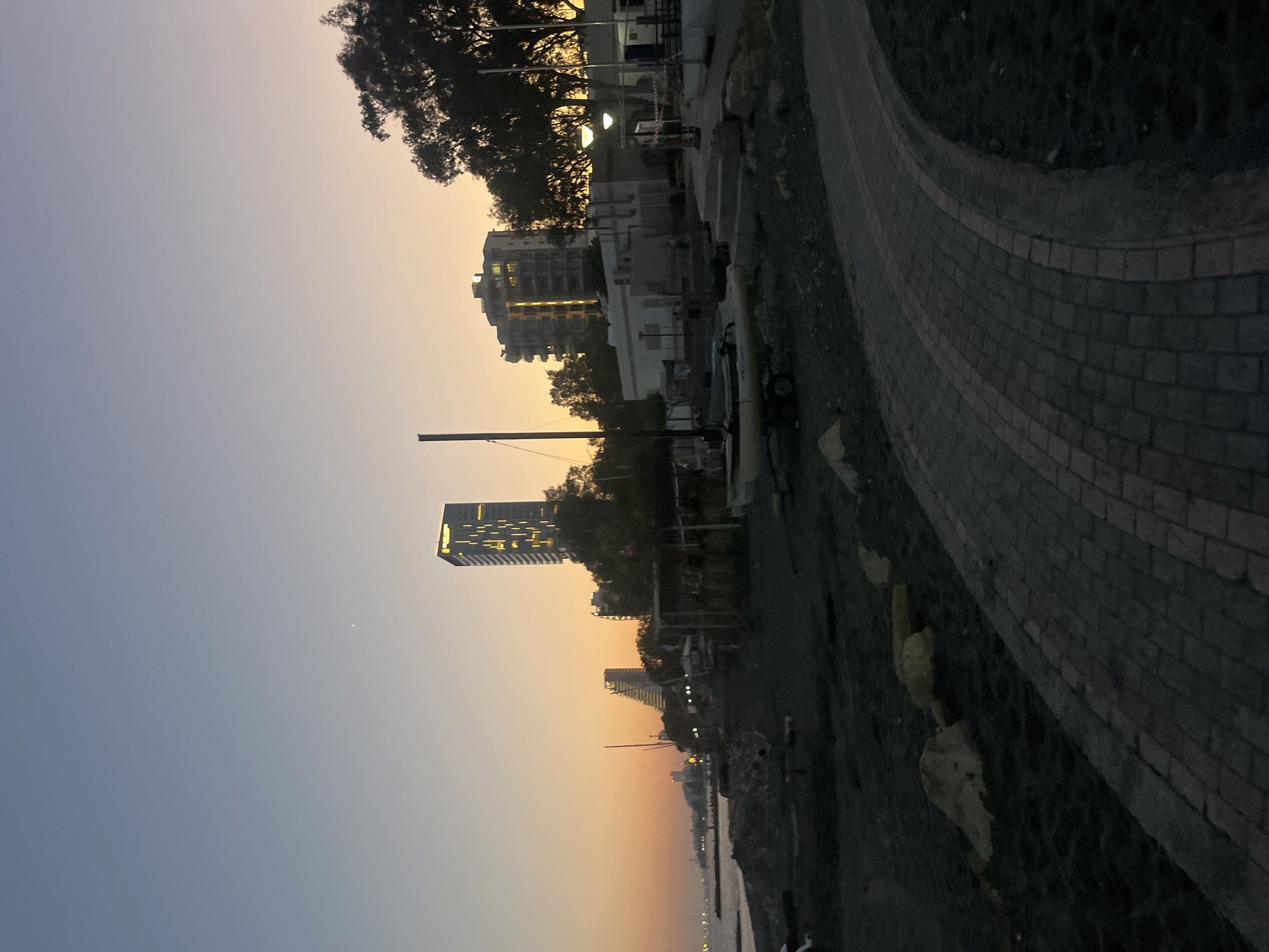 This image depicts a twilight scene on the coast of Agios Tychona Community, Cyprus. The sky displays a gradient of soft blues and oranges, indicative of sunset or sunrise. In the background, modern high-rise buildings are illuminated, with one particularly prominent skyscraper featuring vertical light accents. A cluster of trees, silhouetted against the sky, stands to the right.
The foreground consists of a dark-colored beach or shoreline, with scattered rocks. A paved walkway, composed of interlocking bricks in a curved pattern, leads into the scene from the lower right. On the beach area, there are several objects, including what appears to be a small sailboat on a trailer, and various structures that seem to be sheds or open-air stalls. The lighting is dim, suggesting either early morning or late evening. No people are clearly visible in the frame.