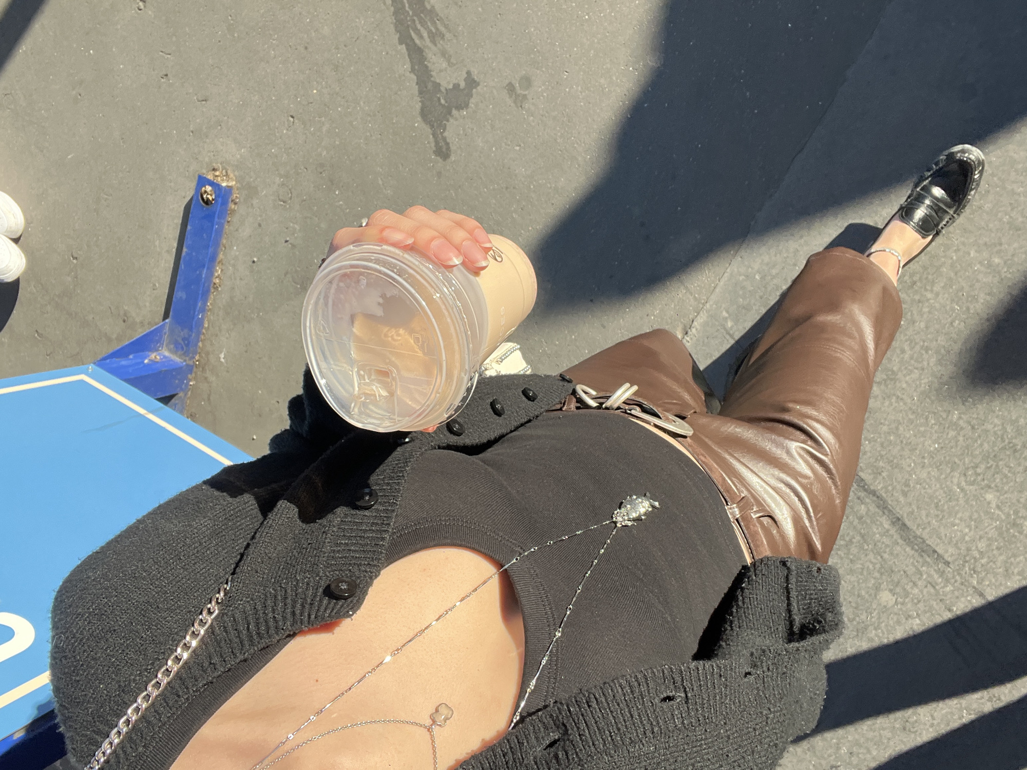 The image captures a top-down, first-person perspective of a person's torso, leg, and hand outdoors on a sunny day in what is contextually described as Paris, France.
The main subject is a person holding an iced coffee in a clear plastic cup. The cup has some visible text, specifically the letters "ES" and a faint logo. The person's hand has manicured nails with a natural finish.
The individual is dressed in a black ribbed knit cardigan, unbuttoned, revealing a black top underneath. They wear brown leather or faux leather pants. A silver belt buckle is visible at the waist. On their left foot, a black loafer shoe with silver detailing is worn, accompanied by a delicate silver anklet. Two layered silver necklaces adorn their neck and chest: one with a small, clear charm, and another with a more elaborate silver pendant.
The setting appears to be an urban environment, with a grey asphalt surface underfoot. Strong shadows are cast on the ground, indicating bright sunshine and clear weather, likely during the mid-day or afternoon. Part of a blue metal structure, possibly a bench or stand, is visible on the left, with a partial white stripe on its side. No specific location cues directly identifying Paris are visible within the image itself.