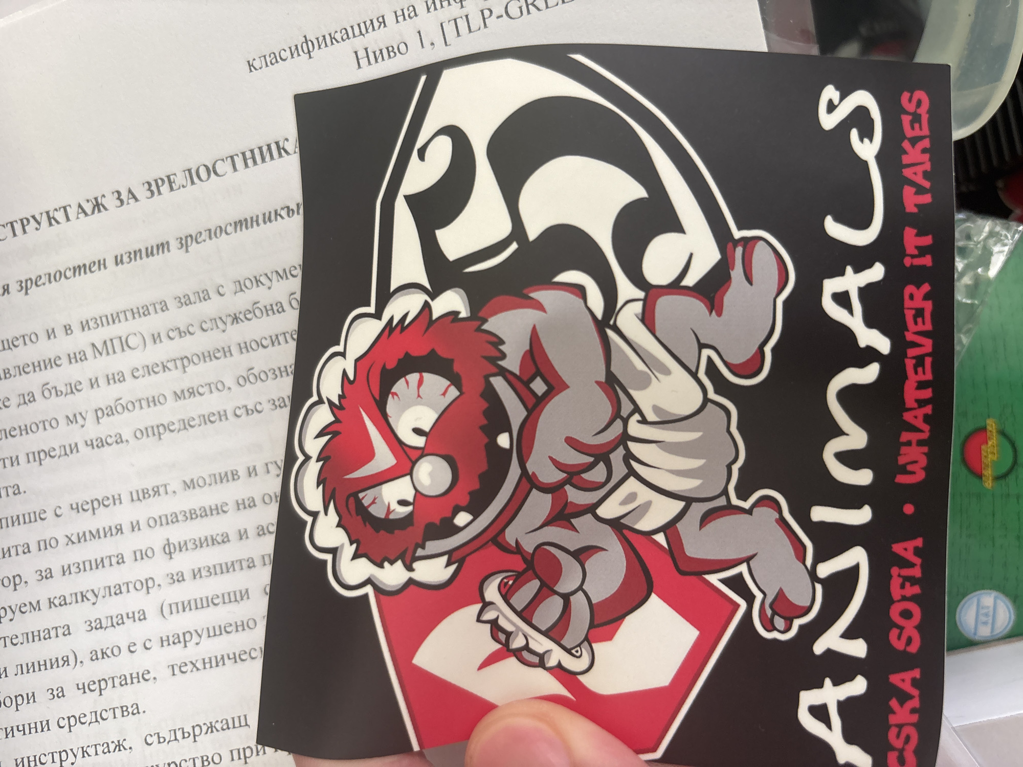 A human hand is holding a multi-colored sticker in front of a white document with black text. The sticker is irregularly shaped, resembling a stylized shield or crest, predominantly black, white, and red.
The central subject of the sticker is a cartoon character, depicted as an aggressive, beast-like creature. It has red, spiky fur/hair, large, bloodshot eyes, sharp teeth, and a grey body. One arm wears a spiked bracelet. The creature is posed dynamically, appearing fierce or determined. Behind the character, a large white and red graphic element is visible, possibly a stylized letter or symbol.
To the right of the character, large white text runs vertically, spelling "ANIMALS". Below this, in smaller red letters, is the phrase "CSKA SOFIA • WHATEVER IT TAKES". CSKA Sofia is a well-known Bulgarian football club.
The document behind the sticker is a white page with text printed in Bulgarian. Visible headings and phrases include "класификация на инф" (classification of inf), "Ниво 1, [TLP-GRE" (Level 1, [TLP-GRE), and "СТРУКТАЖ ЗА ЗРЕЛОСТНИКА" (INSTRUCTION FOR GRADUATE/MATURITY EXAM). The text details instructions for an exam, mentioning aspects like documents, electronic devices, workspace, time, writing materials (black ink, pencil, eraser), subjects (chemistry, environmental protection, physics), calculators, assignments, drawing tools, and technical equipment.
The setting appears to be indoors. In the blurry background to the right, parts of a green container and a clear plastic object are faintly visible. The presence of Bulgarian text on the document and the "CSKA SOFIA" reference strongly suggest the location is Bulgaria.