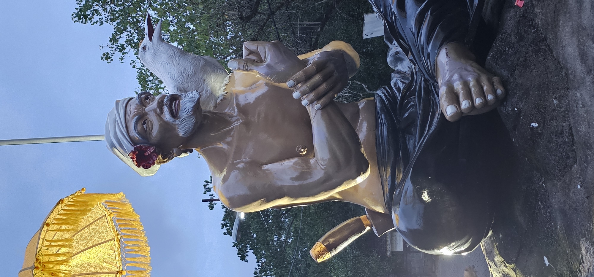 This media file captures a large, multi-figured statue outdoors in Denpasar, Indonesia, illuminated by ambient and artificial light, suggesting either dusk or dawn.
The central and most prominent figure is a man with a light brown skin tone, a white beard, and a calm expression. He wears a turban-like white head covering, from which a red flower emerges near his left ear. A white bird, resembling a dove or pigeon, perches on his right shoulder, looking upwards with its beak open. His right hand is raised, while his left hand rests on his torso.
Intertwined with the central figure are other elements. A pair of hands, with light-colored fingernails, gently touches the central figure's side. Below, a bare foot with light-colored toenails, emerging from dark, draped clothing, rests on a dark, textured surface, possibly a rock. A dark, bulbous object, perhaps a water pot or kamandalu, is situated near the foot. A long, ornate, cylindrical object with a golden tip, possibly a staff or pipe, extends from the lower part of the statue.
In the lower-left corner, a bright golden, fringed, umbrella-like structure (possibly a ceremonial chattra or parasol) is visible, glowing from internal illumination. The background consists of a clear, light blue sky with some green foliage from trees visible at the top. The scene depicts a static, serene moment, likely of religious or spiritual significance. No visible text is present.
