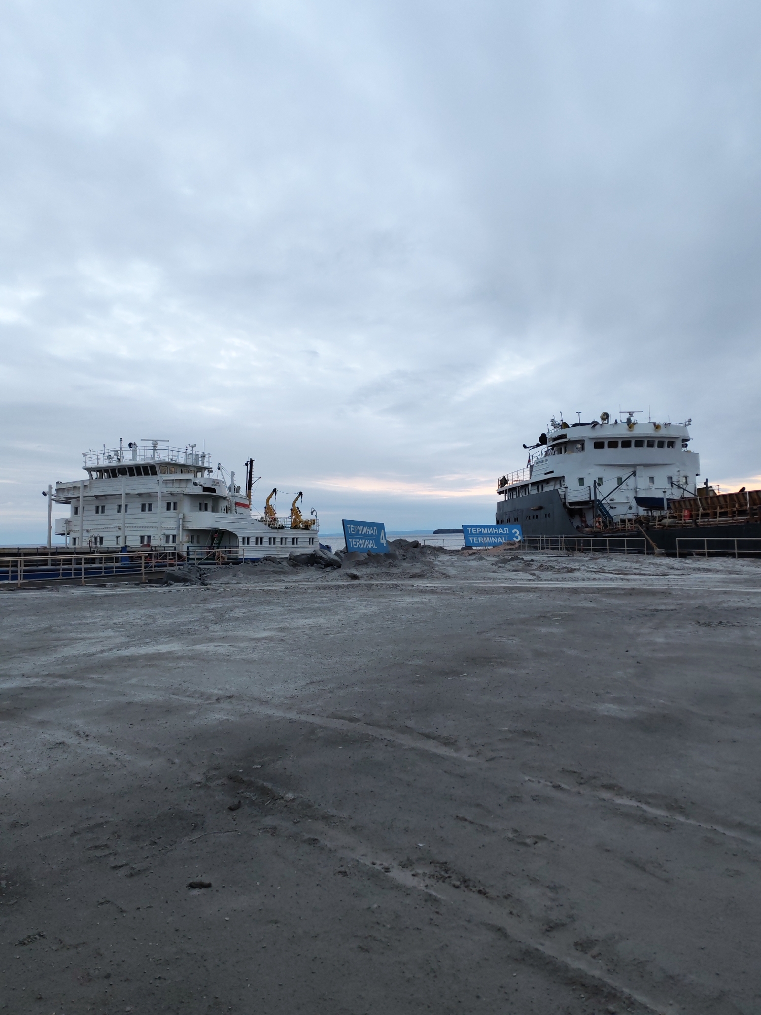 The image depicts a port area in Shcheleiki, Russia, under an overcast sky. Two large vessels are docked at Terminal 3 and Terminal 4, respectively. The ground appears to be covered in a gray, sandy material, possibly cement. The vessels are predominantly white and gray, with visible cranes on the vessel docked at Terminal 4. The signs indicating the terminals are blue and white. The scene is still, lacking apparent human activity. The image suggests an industrial or commercial port environment.