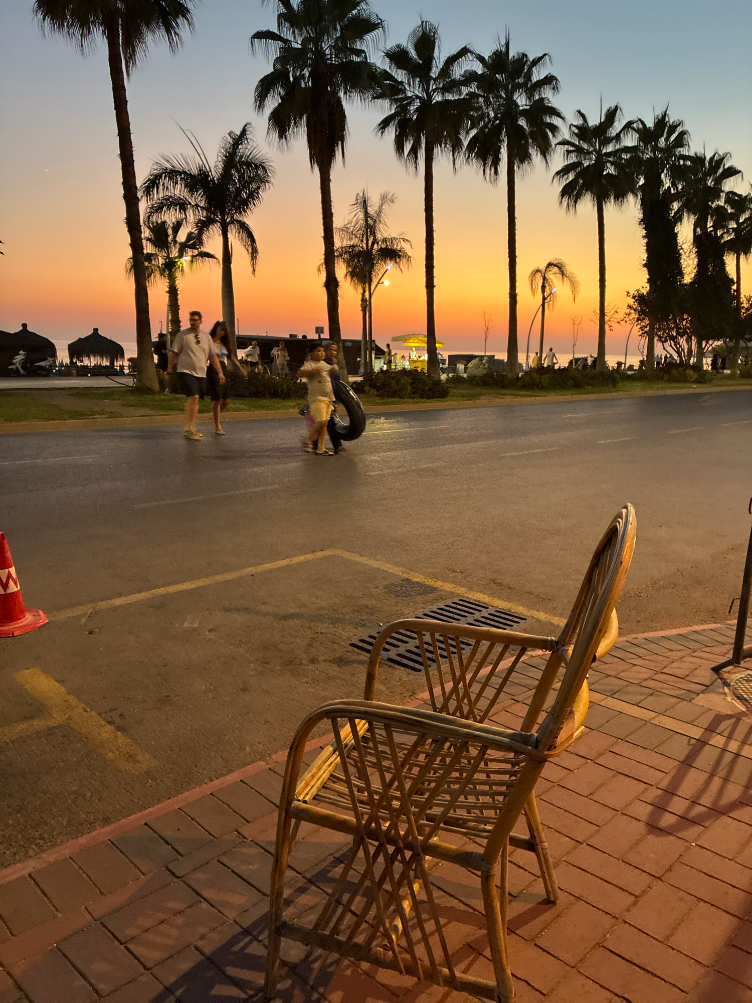 The image depicts a sunset scene in Alanya, Turkey, likely on or near Atatürk Boulevard, a prominent coastal road. In the foreground, a light-colored wicker chair sits on a red brick sidewalk, casting a shadow on the ground. A street grate is visible to the left of the chair, and a traffic cone with an orange and white pattern is in the lower left corner of the frame.
The middle ground shows a paved road with yellow lane markings. Several people are walking on the sidewalk across the street. Among them, a couple is walking towards the left, with a man in a light-colored shirt and shorts, and a woman in a dark dress. Further to the right, a person is carrying a large black tire. Behind the sidewalk, a row of tall palm trees silhouetted against the sky dominates the background. Beyond the trees, the calm sea is visible, reflecting the warm hues of the sunset. The sky transitions from a soft blue at the top to vibrant shades of orange and yellow near the horizon. Distant structures and lights suggest a populated area.