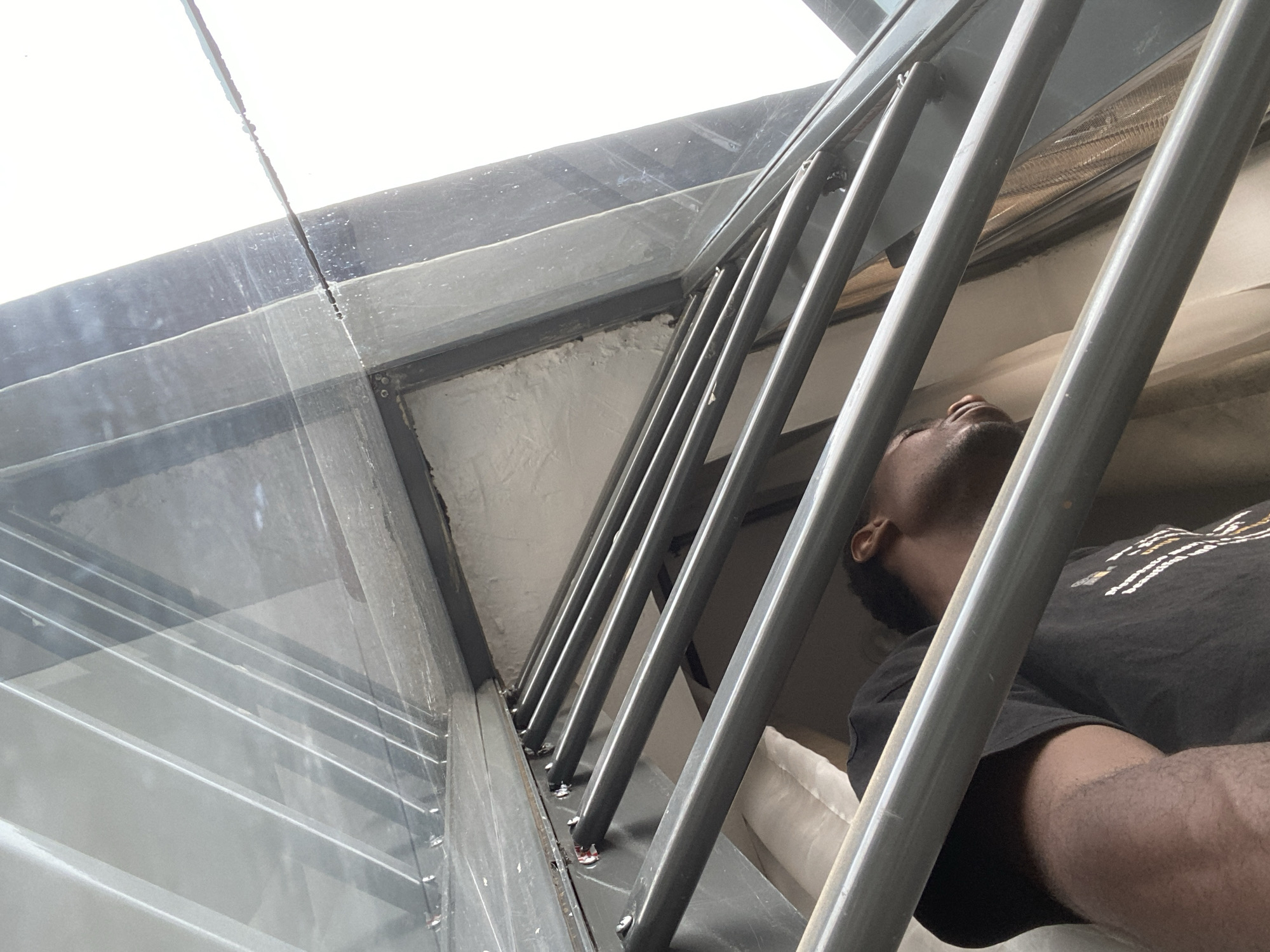 The image shows a man lying down inside a building in Ibadan, Nigeria. The man is Black and is wearing a black t-shirt. He is lying on a white cushion. The room has a metal staircase. The windows are clear glass. Rain is falling on the window.