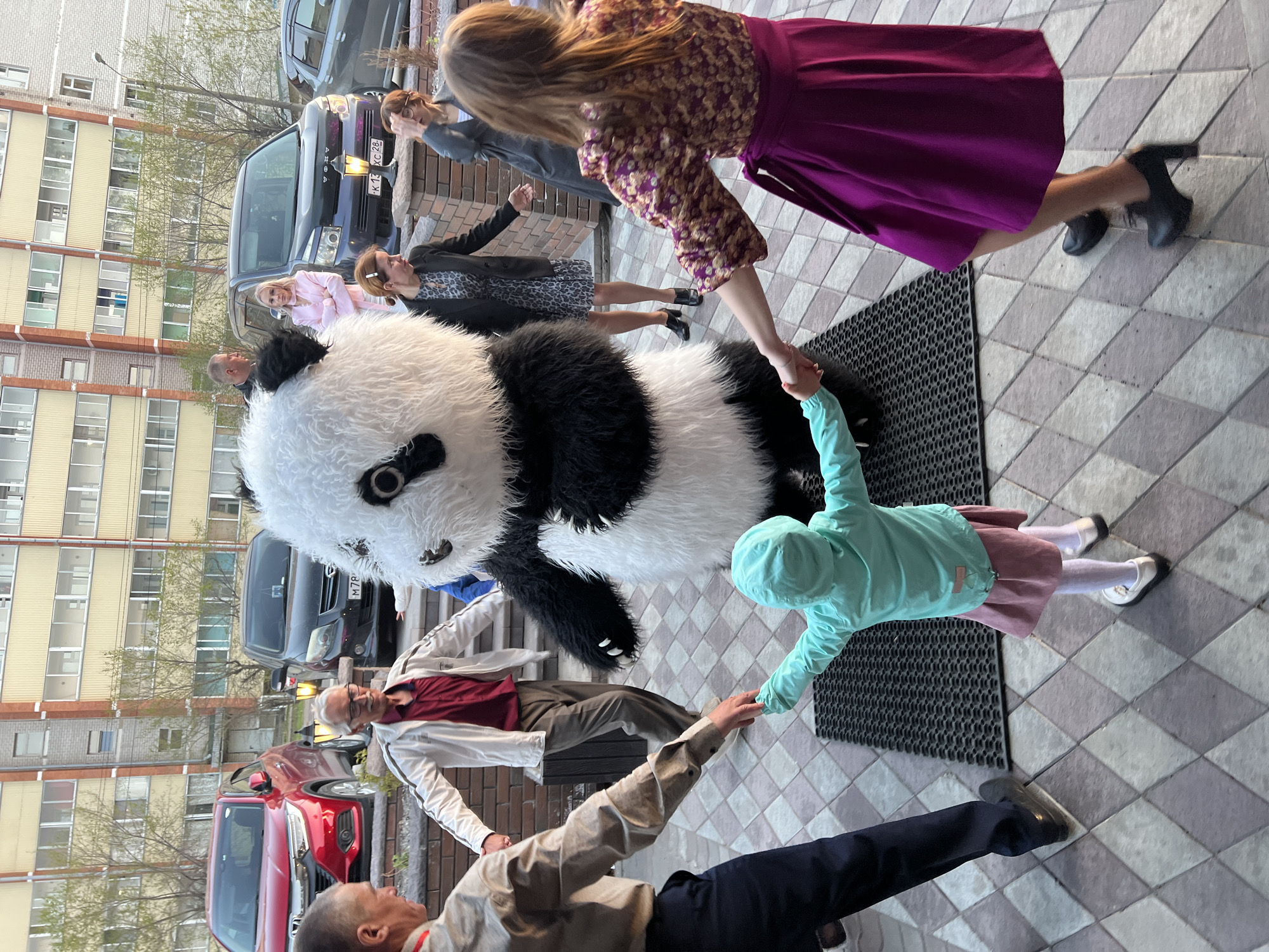 Outside a multi-story building of Unknown, Unknown, people have joined hands in a ring around an actor dressed in a panda suit. The group consists of a child, at least five women, and at least four men. Behind them are parked cars, of which at least one is red, another is dark grey, and another is black. A black rubber mat is also on the floor behind the panda. The overall mood of the scene seems happy.