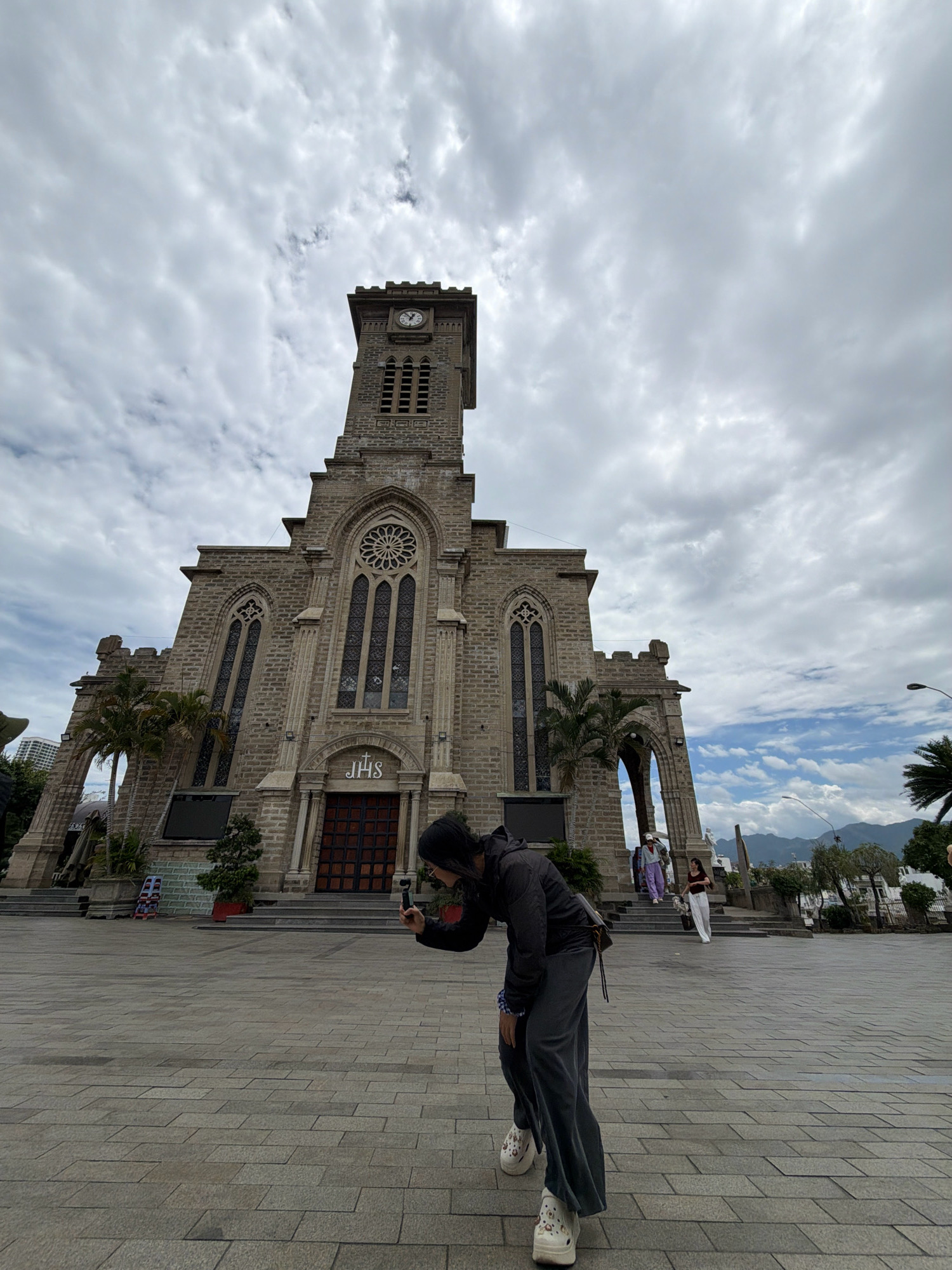 A person in the foreground, viewed from the chest down, is holding a smartphone and appears to be filming or taking a picture of a large, stone church with a tall bell tower in the background. The person is wearing a dark hooded jacket and loose-fitting dark pants, with white patterned shoes. They are standing on a paved plaza. The church is constructed of light-colored stone, featuring gothic architectural elements like pointed arches and stained-glass windows. A clock is visible on the bell tower. The sky is cloudy with patches of blue. Several other people are visible walking on the steps of the church in the background. This is the Nha Trang Cathedral in Nha Trang, Vietnam. The cathedral is a prominent landmark in the city, known for its French Gothic architecture.