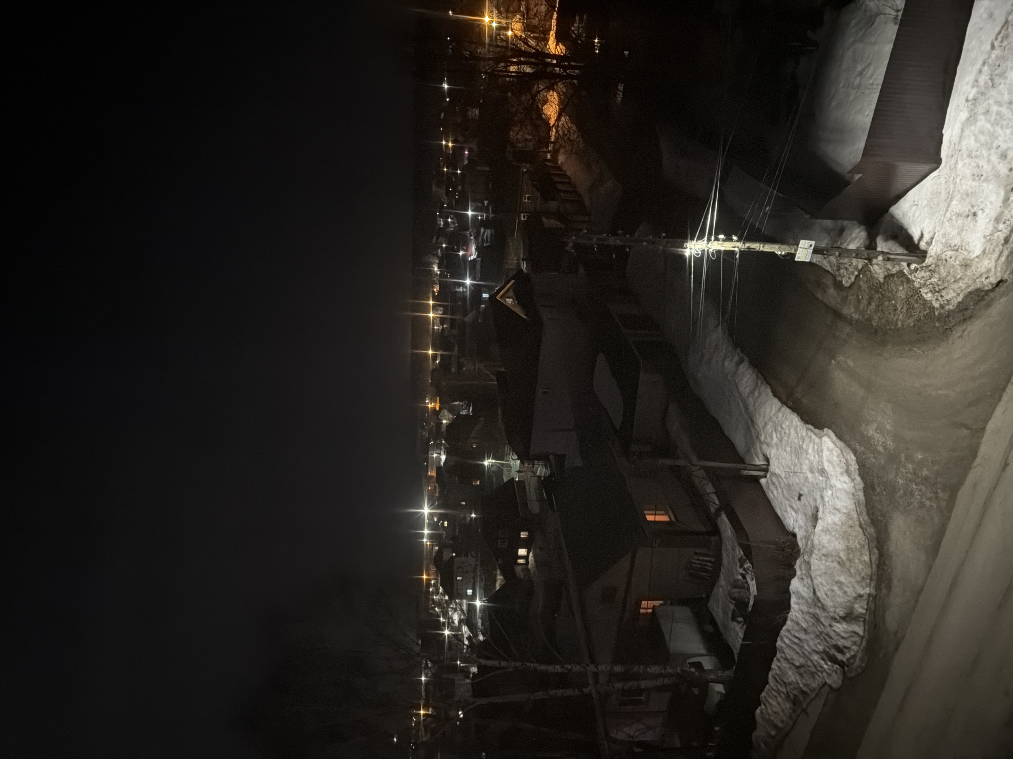 The image shows a night view of Berezniki, Russia. A row of streetlights illuminates the city buildings and streets. Snow is present on the ground and rooftops, particularly on the right side. Power lines run along the street, and a sign is visible on a pole. The left side of the image is dominated by the dark night sky.