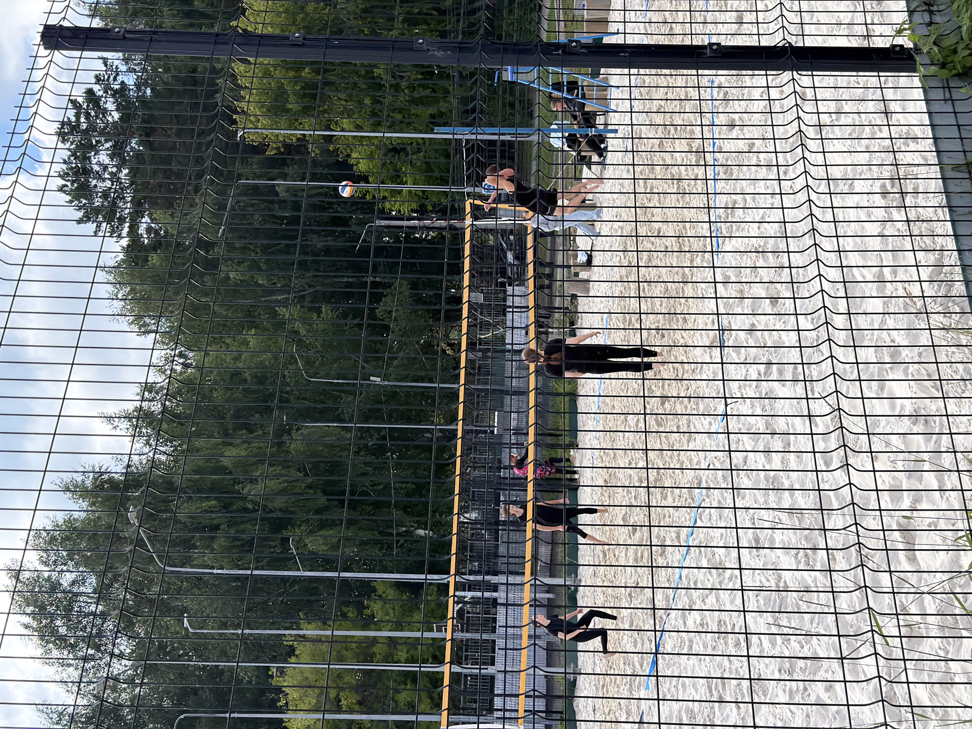 The image shows a beach volleyball court with a game in progress. Four women in black athletic wear are playing on the sand. One woman is serving the ball, another is jumping to block it, and two others are in defensive positions. A volleyball is in the air. The court is enclosed by a black metal fence, and trees are visible in the background. The scene appears to be in a park or recreational area in Chaikovsky, Russia, given the characteristic fencing and the presence of lamp posts common in Russian urban parks.