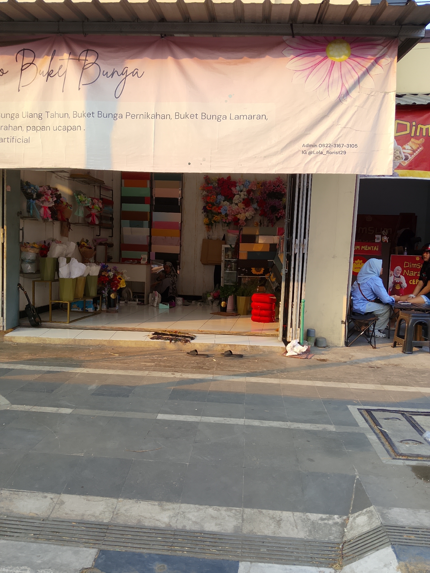 The image shows a flower shop named "Bukit Bunga" in Sambong, Indonesia. The shop displays various flowers and bouquets, including artificial ones. A banner on the shop entrance advertises services like birthday bouquets, wedding bouquets, and engagement bouquets. The shop also provides contact information, including a phone number and an Instagram handle. Inside the shop, a woman is seen sitting at a table, possibly interacting with a customer. The storefront is open, revealing a display of flowers and a seating area. The exterior of the shop is paved with gray tiles, and there are some potted plants and decorations. A guitar is leaning against a shelf inside the shop.