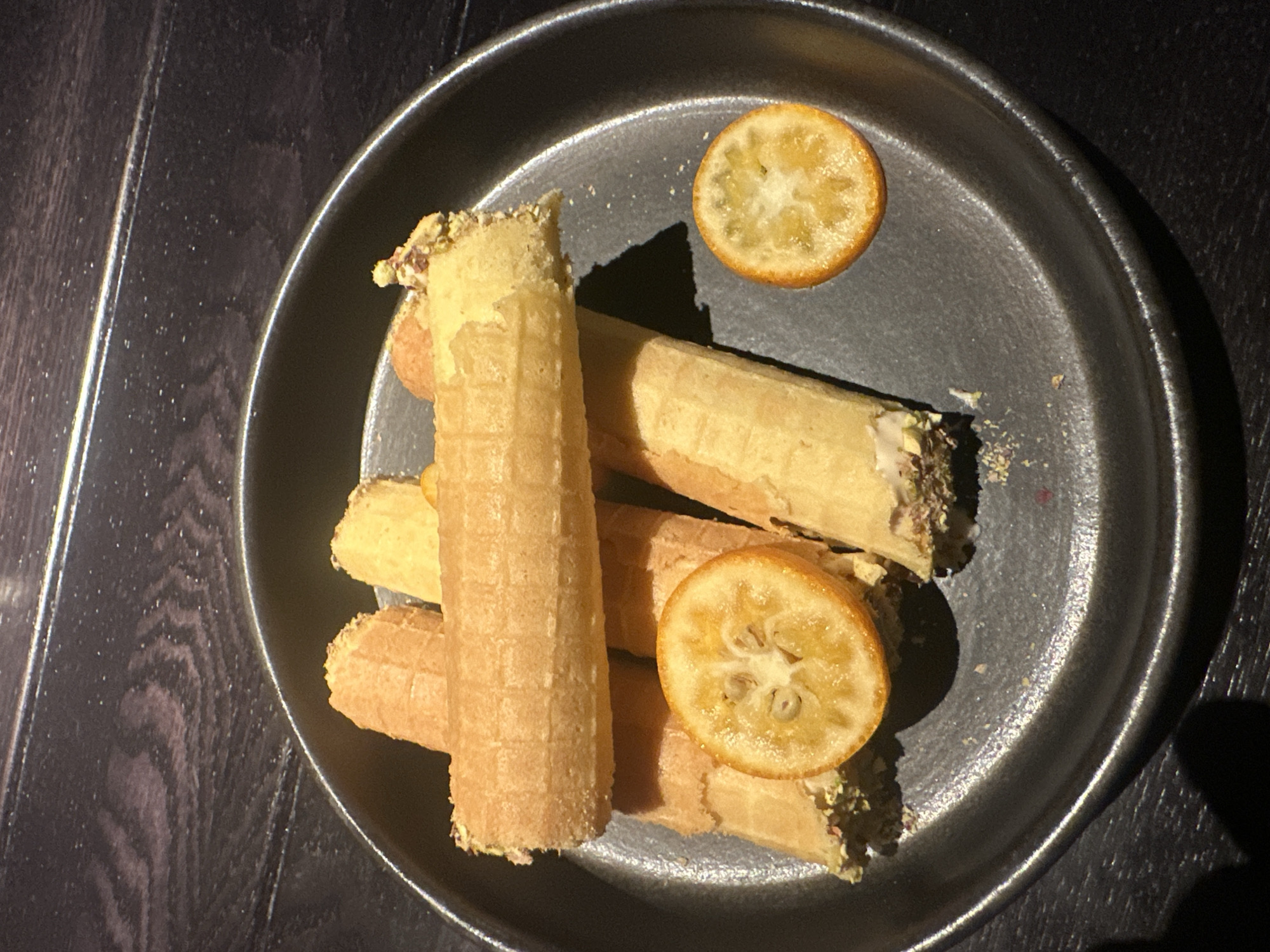 A close-up shot depicts a dark gray plate containing several waffle tubes and two slices of citrus fruit, possibly kumquats. The waffles appear to be filled with a lighter-colored filling, some garnished with green nuts, possibly pistachios. The scene is illuminated by an overhead light source, casting shadows. The plate sits on a dark wooden surface. The location, Saratov, Russia, is not discernible from the image alone.