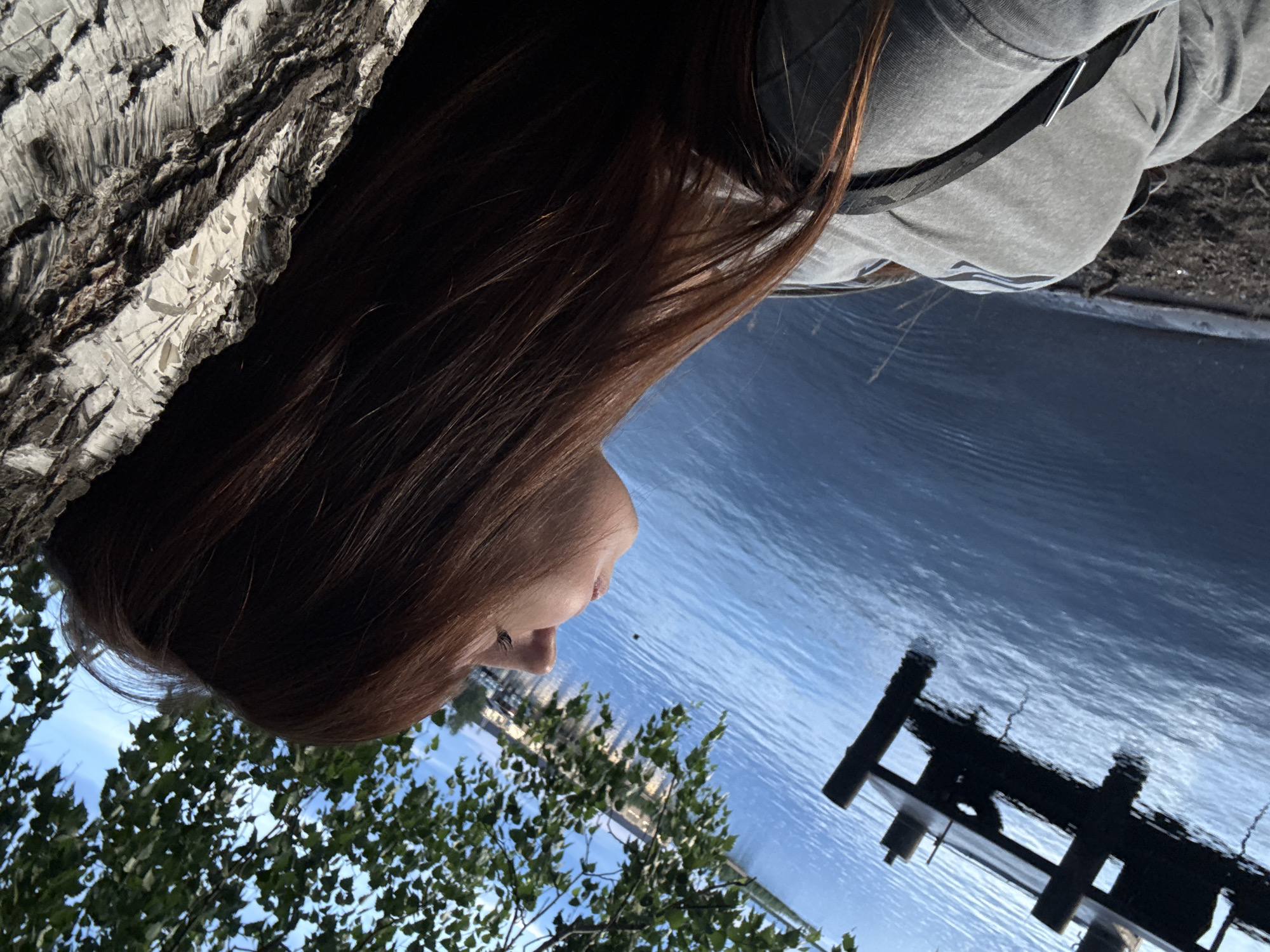 The image shows a person's head and shoulders from a close-up, low-angle perspective. The person has long, reddish-brown hair and is looking downwards and to the right. They are wearing a grey sweatshirt. The background is a body of water with ripples, reflecting the sky and some trees. To the left, a portion of a tree trunk with rough, grey bark is visible. The scene appears to be outdoors, possibly near a lake or river, with foliage from trees framing the bottom left corner. The lighting suggests it is daytime, with sunlight reflecting off the water. The overall mood is relaxed and contemplative. There is no visible text in the image.