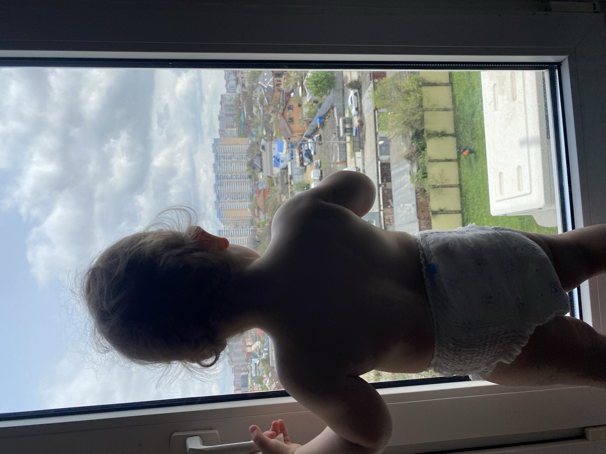 A child wearing a diaper stands at a window, looking out at a cityscape of Unknown, Unknown. The window frame is white. The child's back is towards the viewer. Outside, there is a mix of high-rise apartment buildings, houses, trees, and a patch of grass. The sky is partly cloudy.